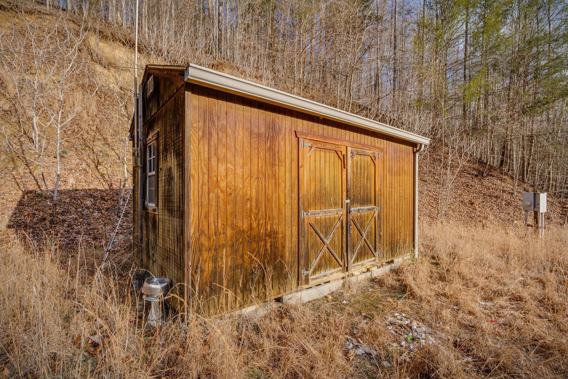 Wooden shed with double doors, set on a hillside. Dry grass surrounds it, trees in the background.