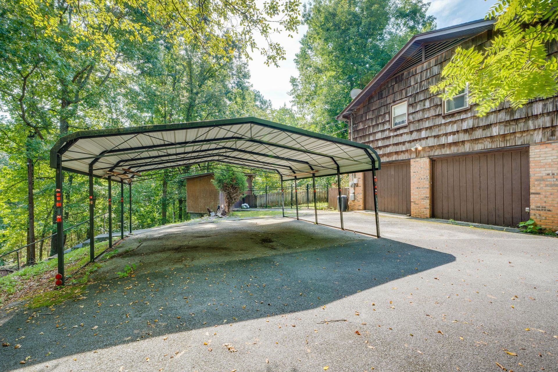 Large metal carport beside a rustic barn, surrounded by trees and gravel driveway