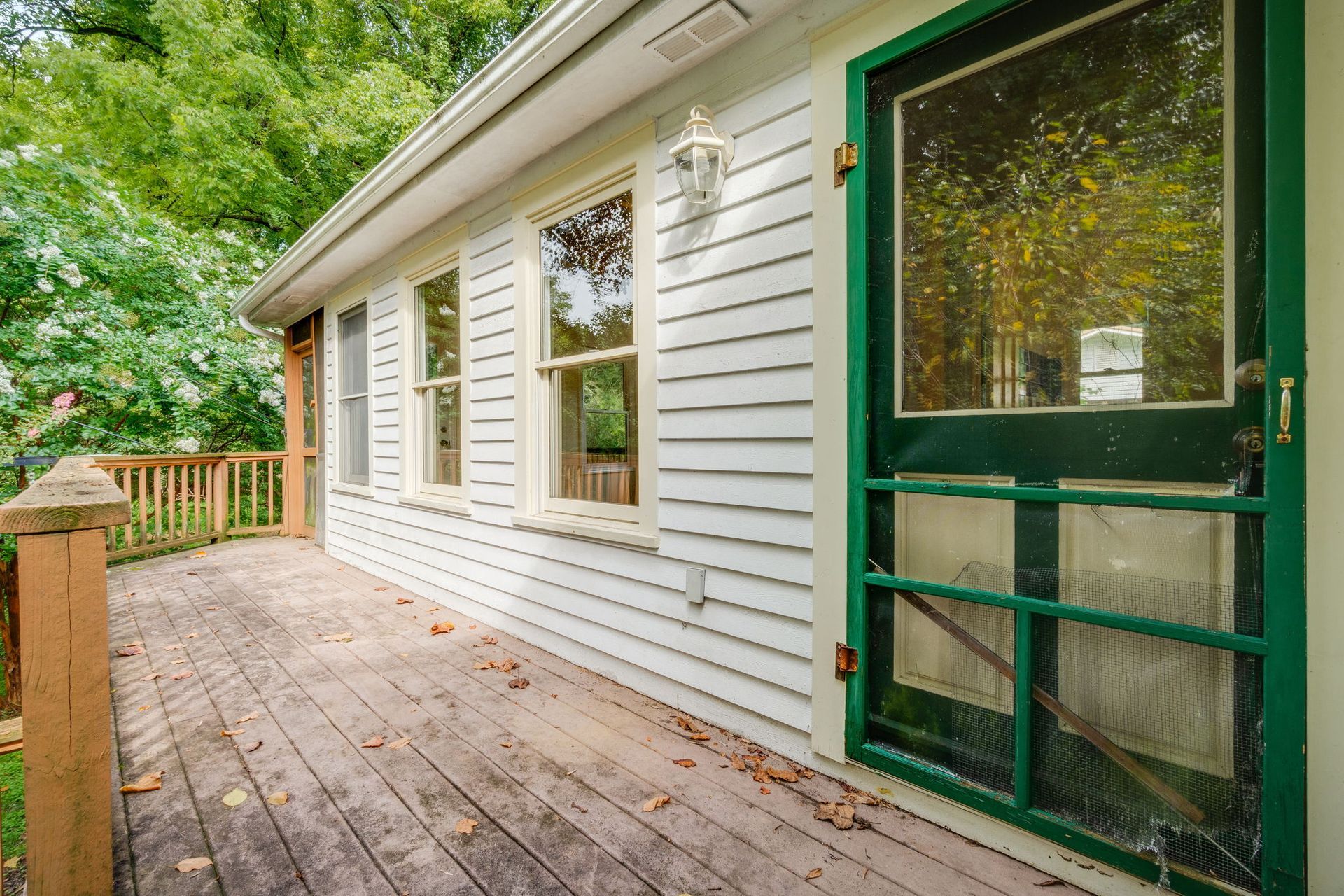 Wooden deck beside a white house with green-trimmed screen door and trees beyond the railing