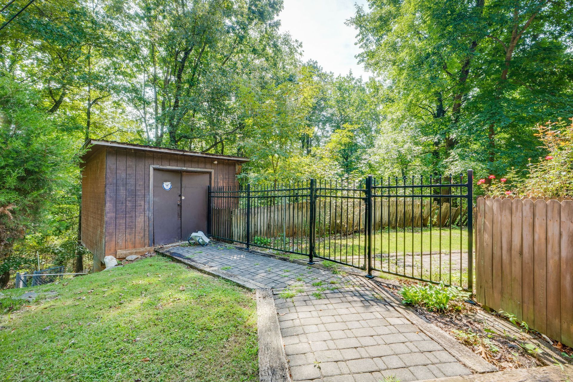 Backyard path leading to a small shed and black metal gate beside a wooden fence and trees