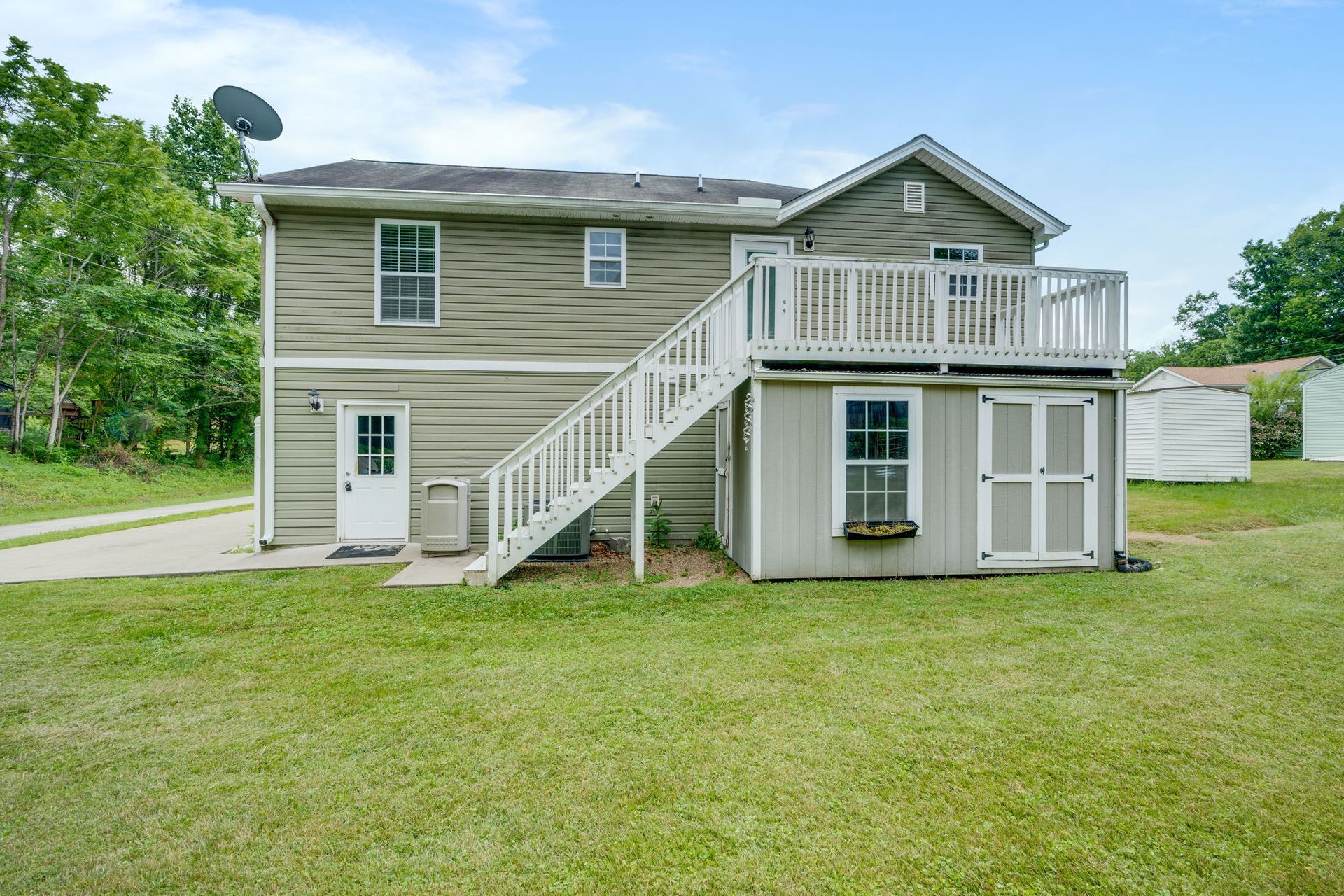 Two-story beige house with white stairs and balcony, set on a grassy yard with trees nearby
