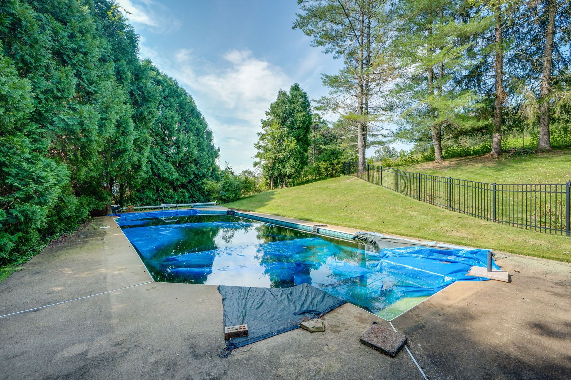 Covered outdoor pool with a bright blue tarp, surrounded by trees and a fence in a sunny yard
