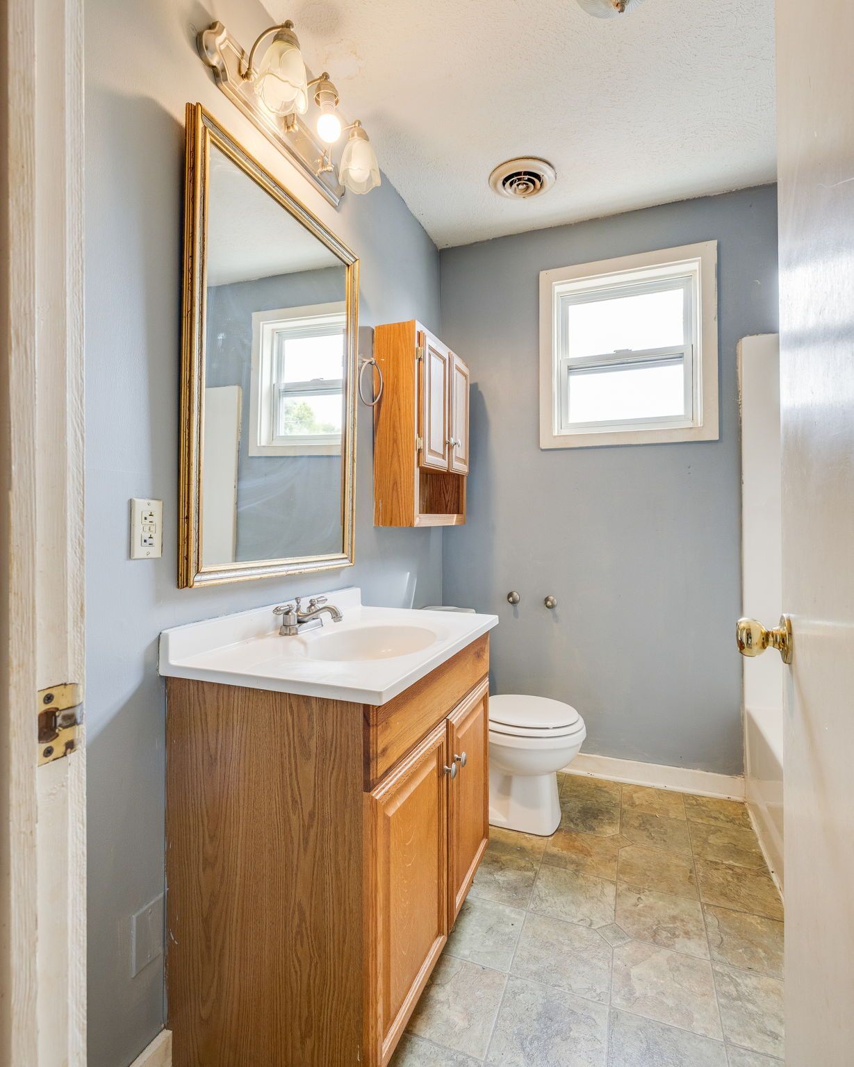 Small bathroom with wood vanity, mirror, toilet, window, and light blue walls