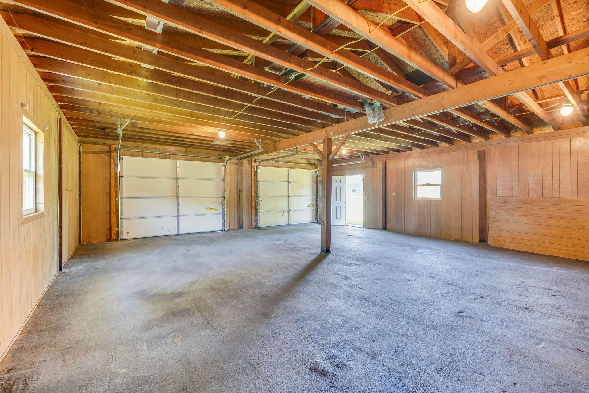 Empty unfinished garage interior with exposed wooden beams, concrete floor, and bright daylight from windows and garage door.