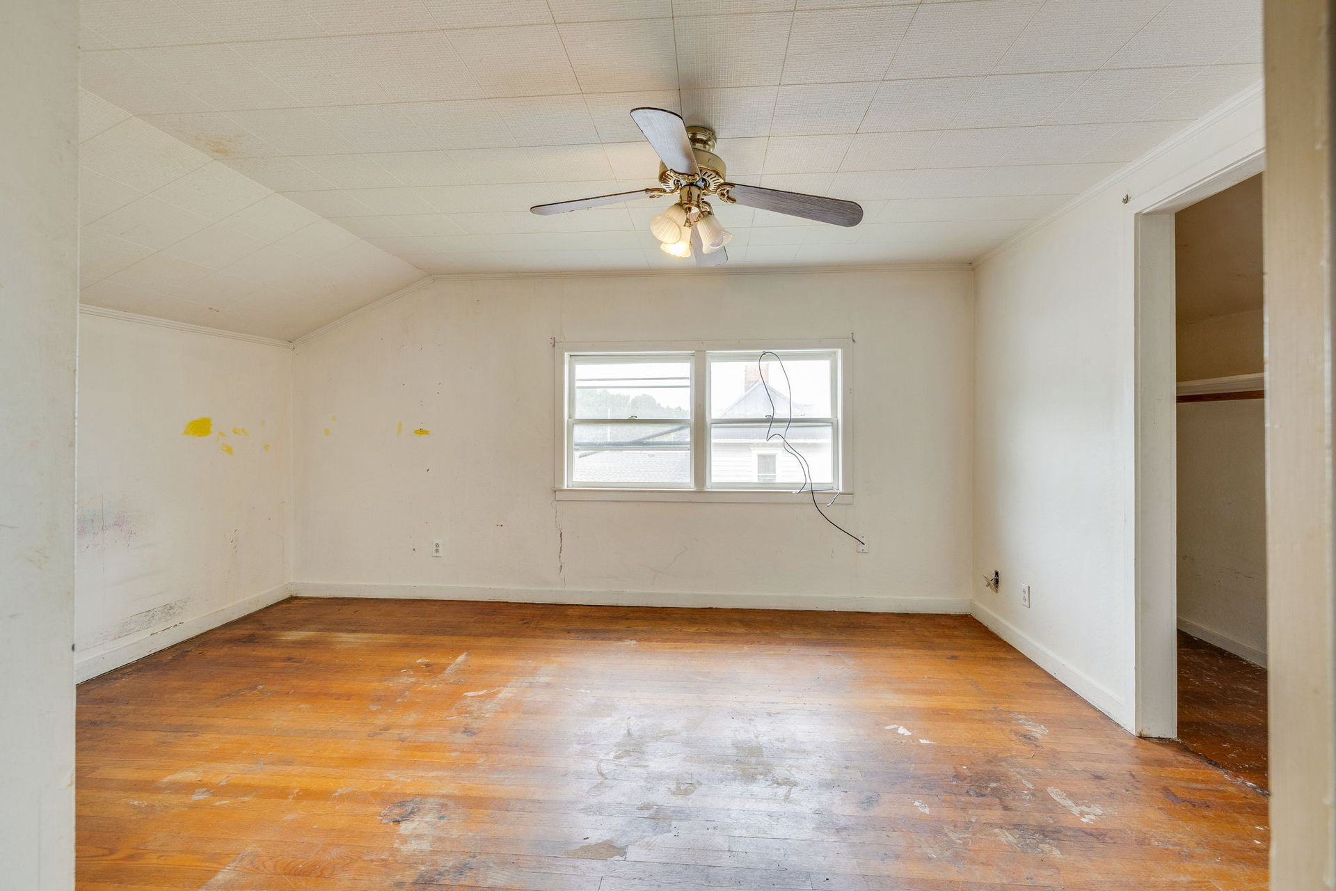 Empty white room with worn hardwood floors, ceiling fan, window, and open closet niche