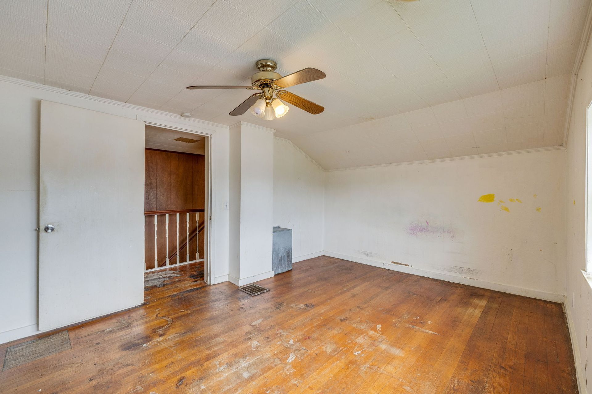 Empty worn room with hardwood floors, white walls, and a ceiling fan, viewed toward an open doorway