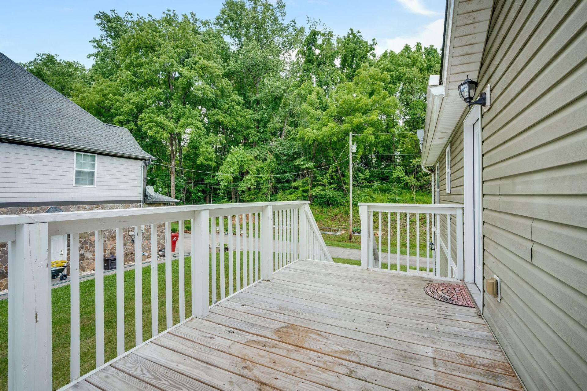 Wooden backyard deck with white railing beside beige house, overlooking green trees and neighboring roof.