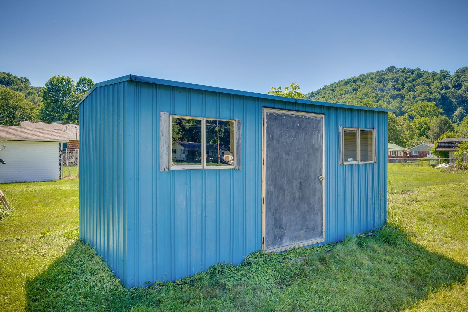 Blue metal shed with two windows and a gray door sitting on a grassy yard