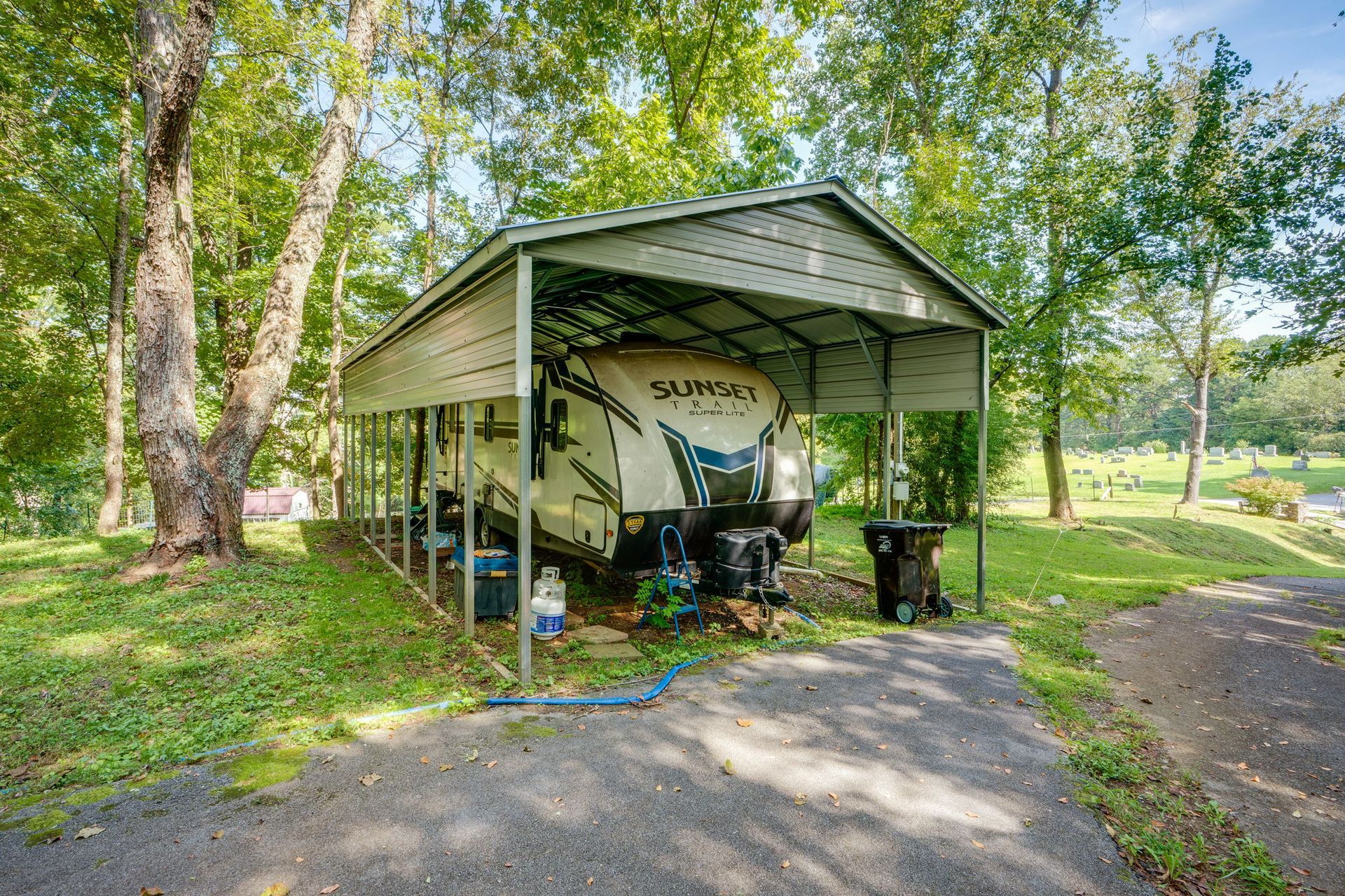 Covered RV parked under a metal carport in a grassy campground with trees and a paved drive