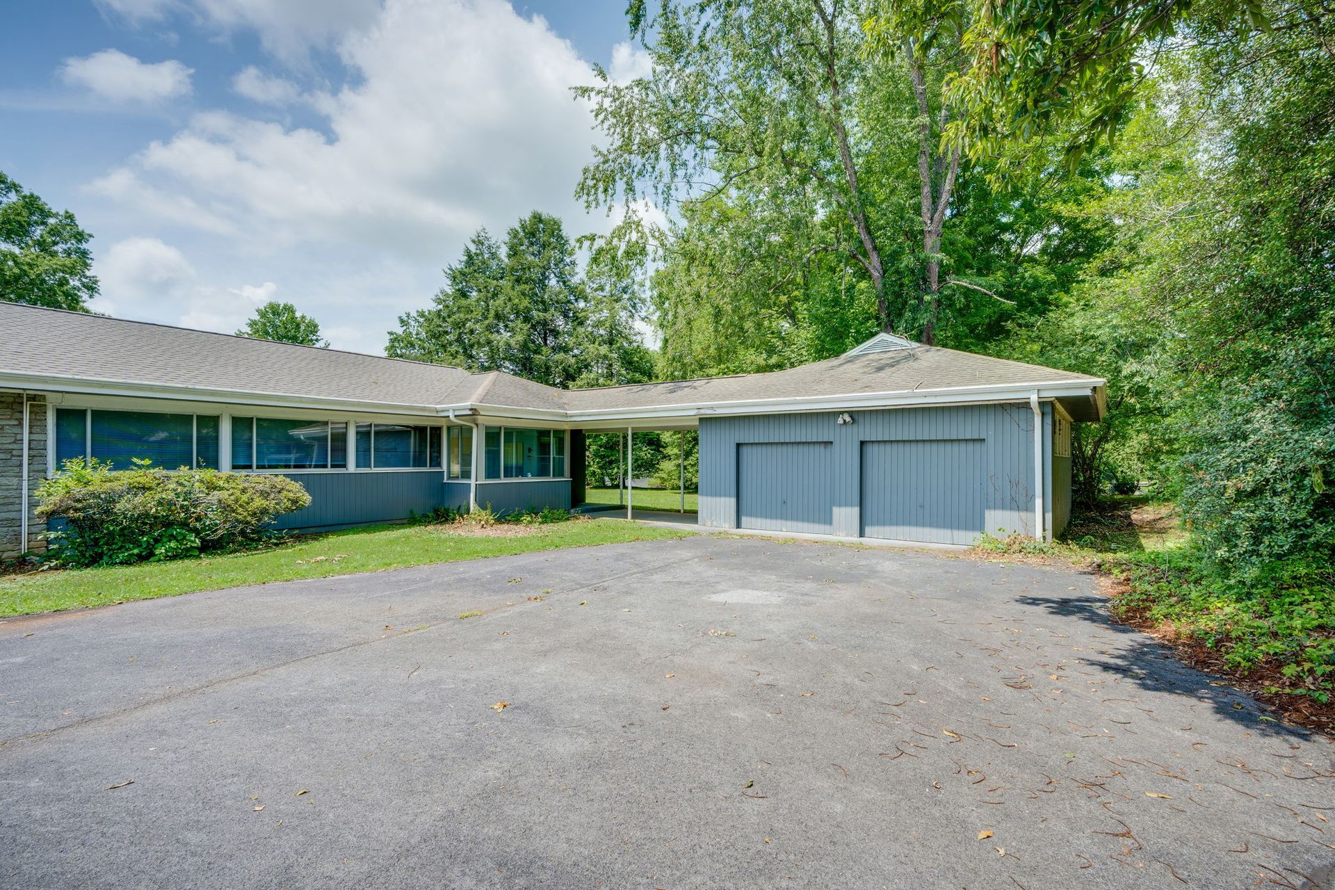 Single-story blue house with a two-car garage and a large gravel driveway, surrounded by trees.