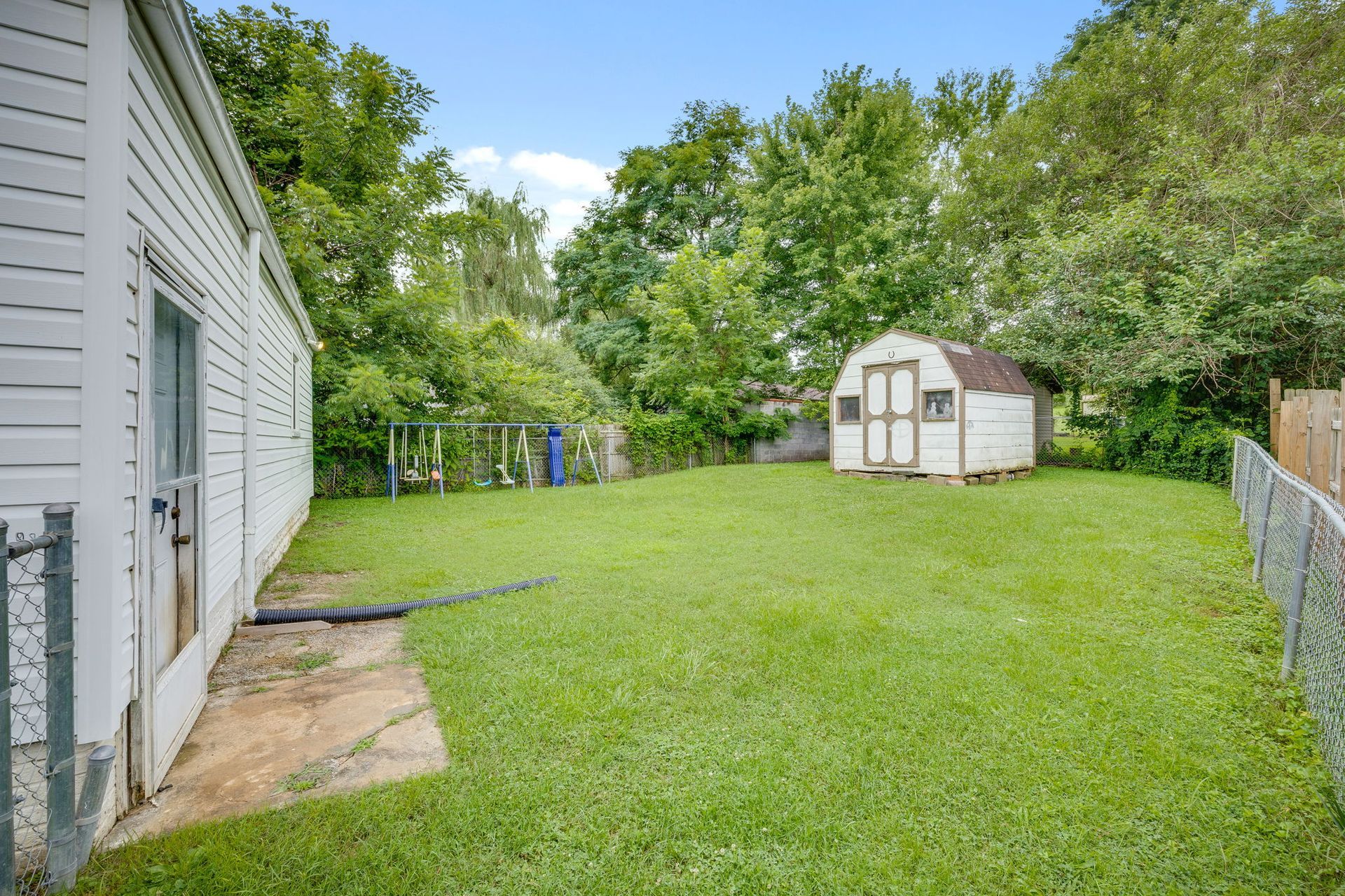 Backyard with green lawn, white house, chain-link fence, trees, and a small shed at the far end