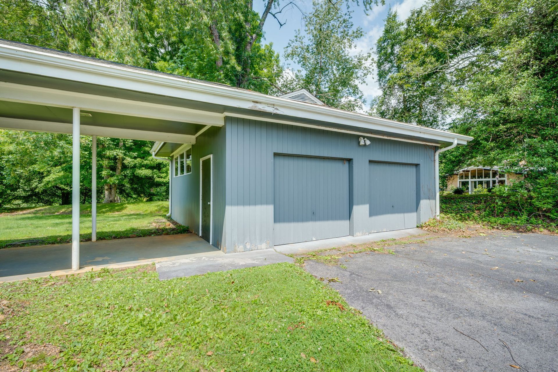 Gray detached garage with carport, surrounded by trees and grass.
