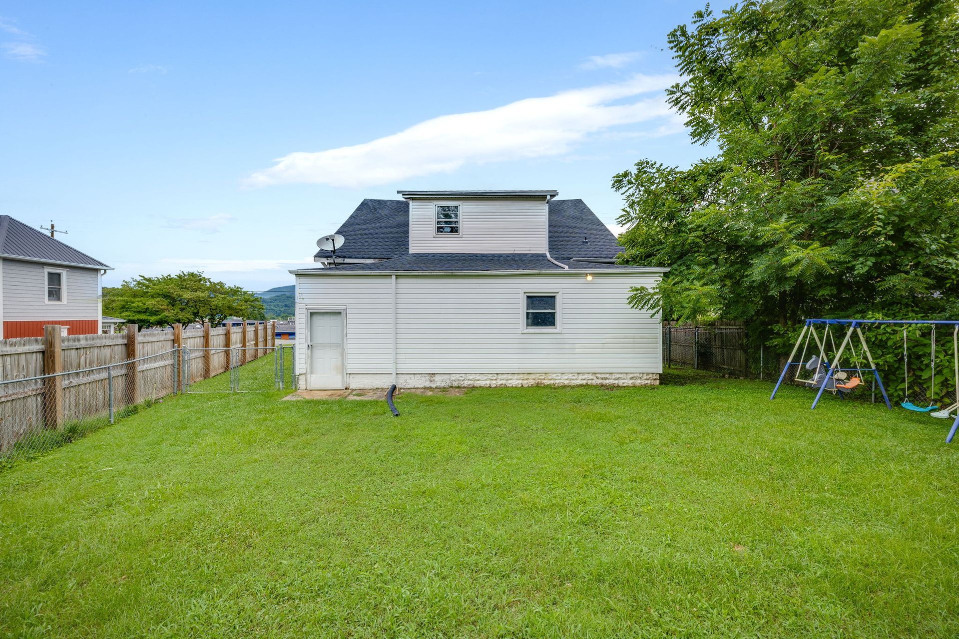 Backyard with a white house, green lawn, wooden fence, and a swing set by trees