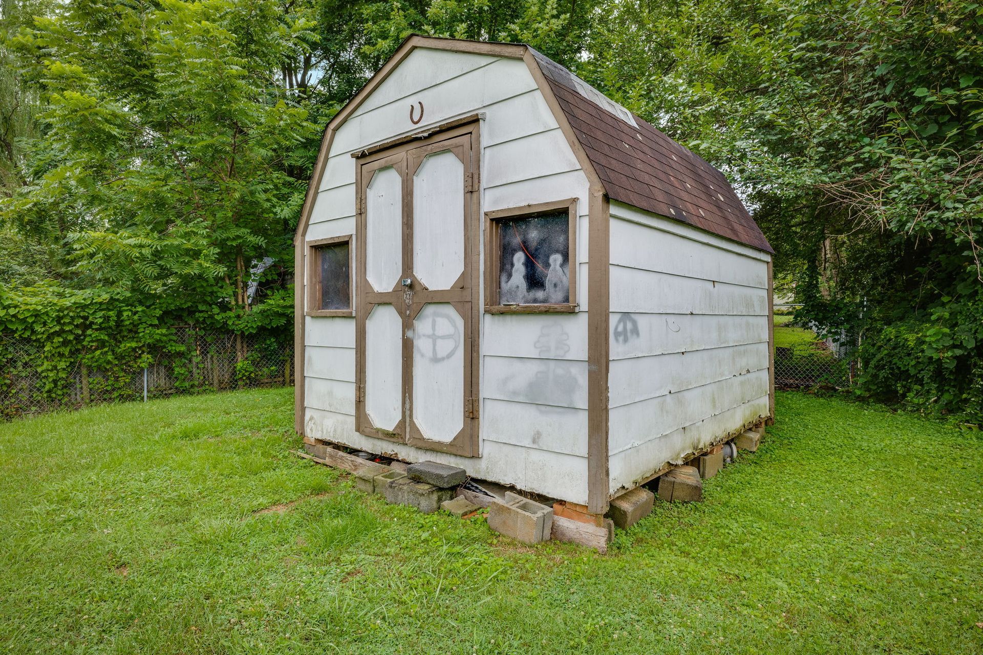 Small white shed with brown roof in a grassy yard, surrounded by trees
