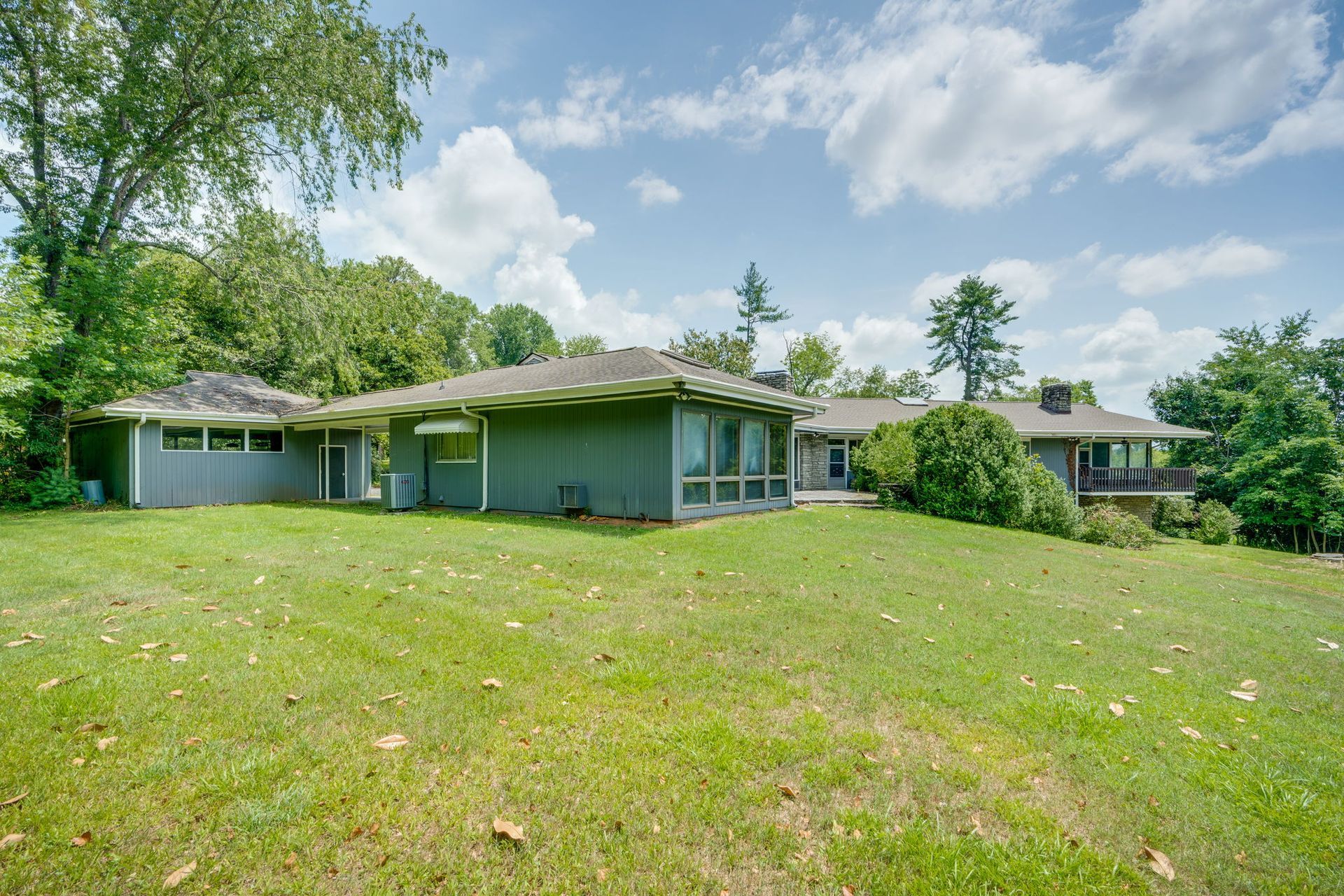 Single-story green house with large lawn and trees under a partly cloudy sky