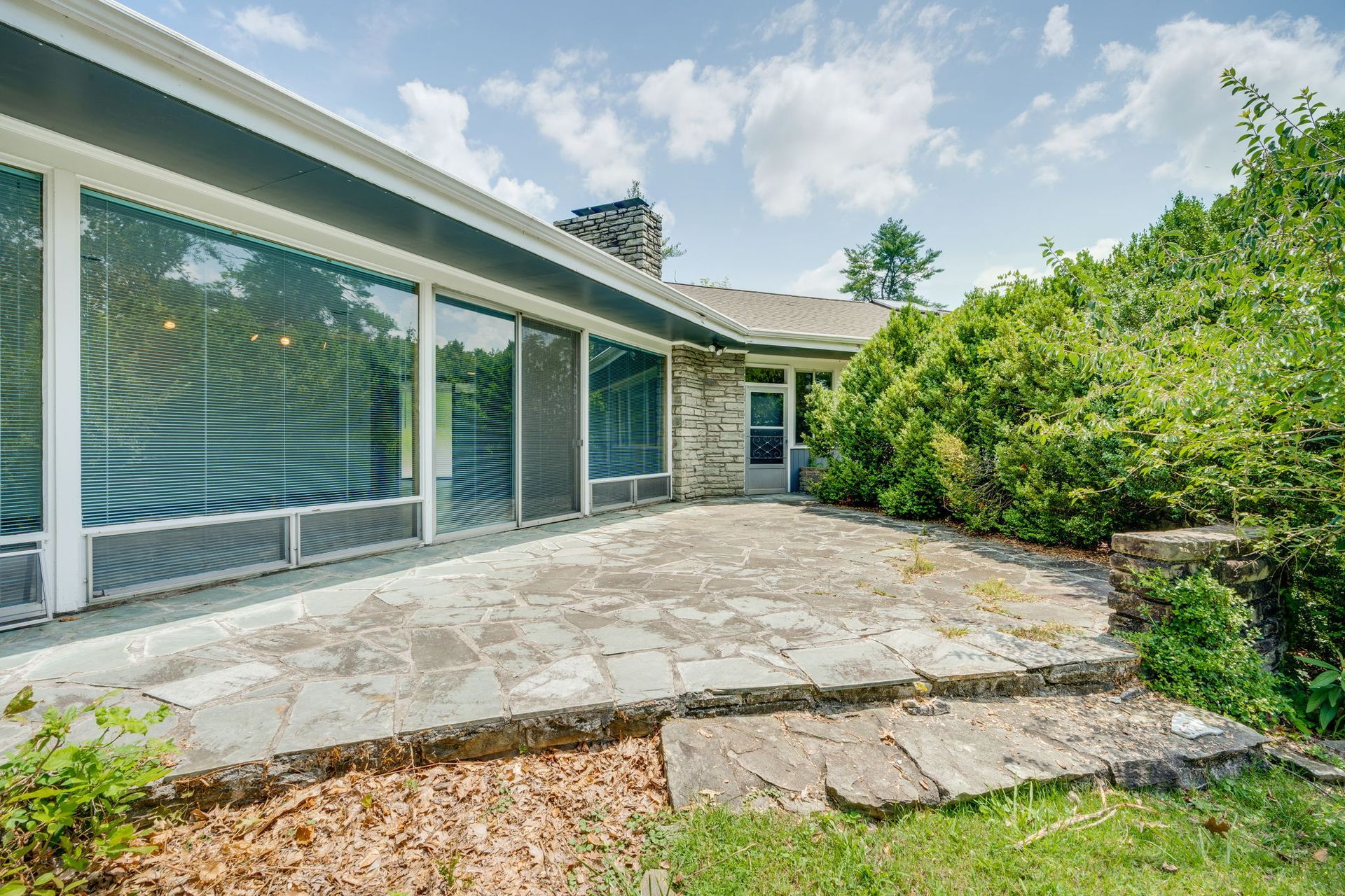 Patio with stone pavers beside a modern house, large windows, and a lush green hedge