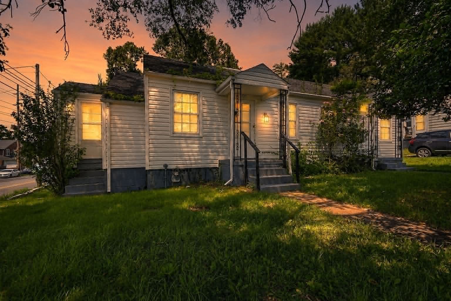 Single-story house with lit windows at dusk, surrounded by trees and a green lawn.