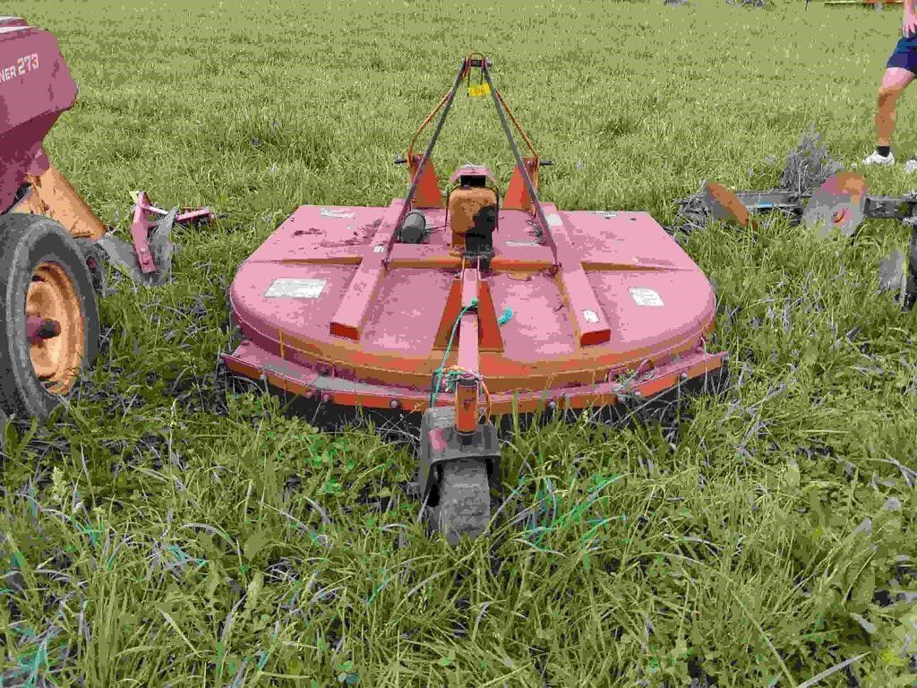 Red rotary mower cutting tall grass in a field, with farm equipment visible at the sides