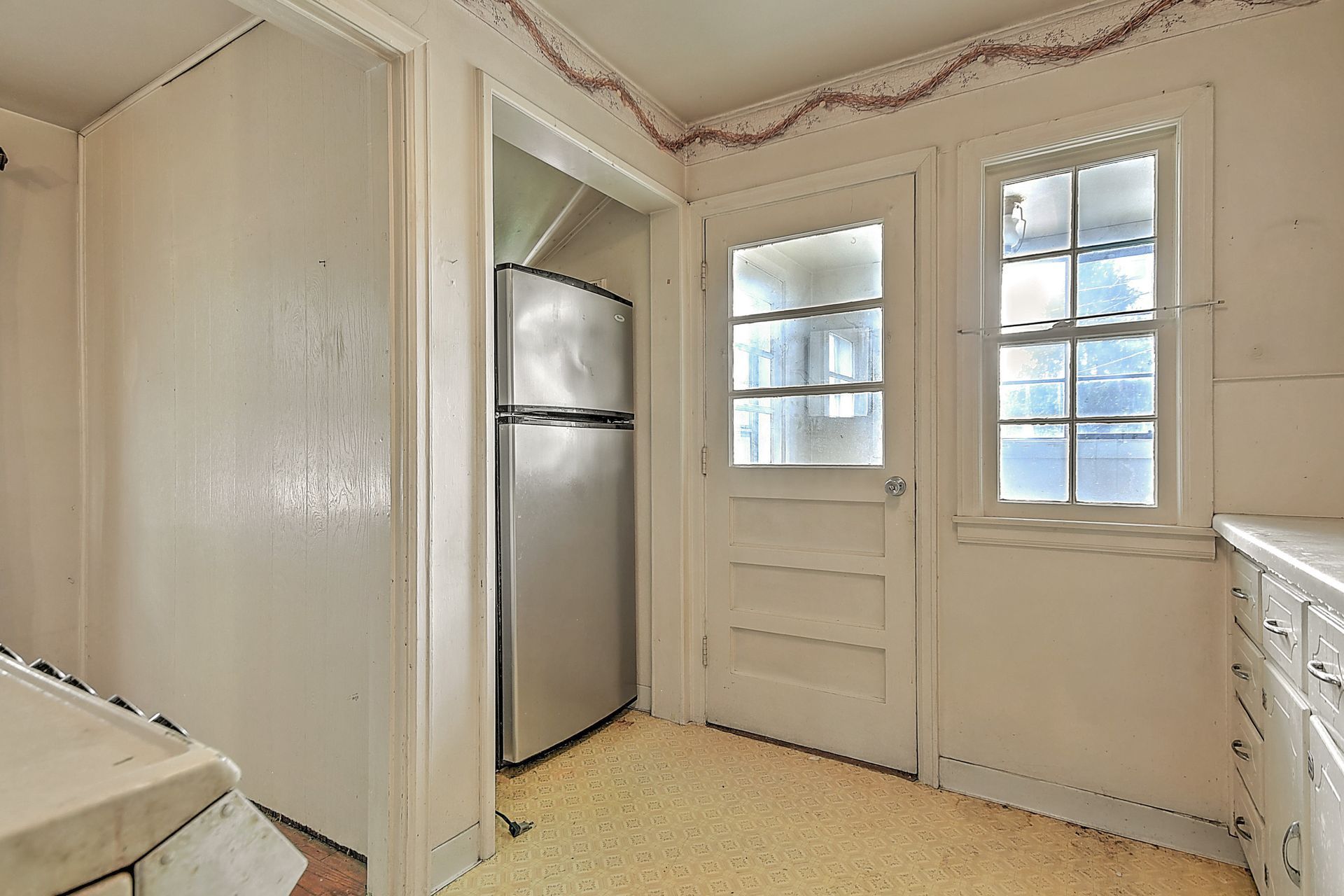 Small kitchen corner with a refrigerator, white door, two windows, and a cabinet under a decorative garland.