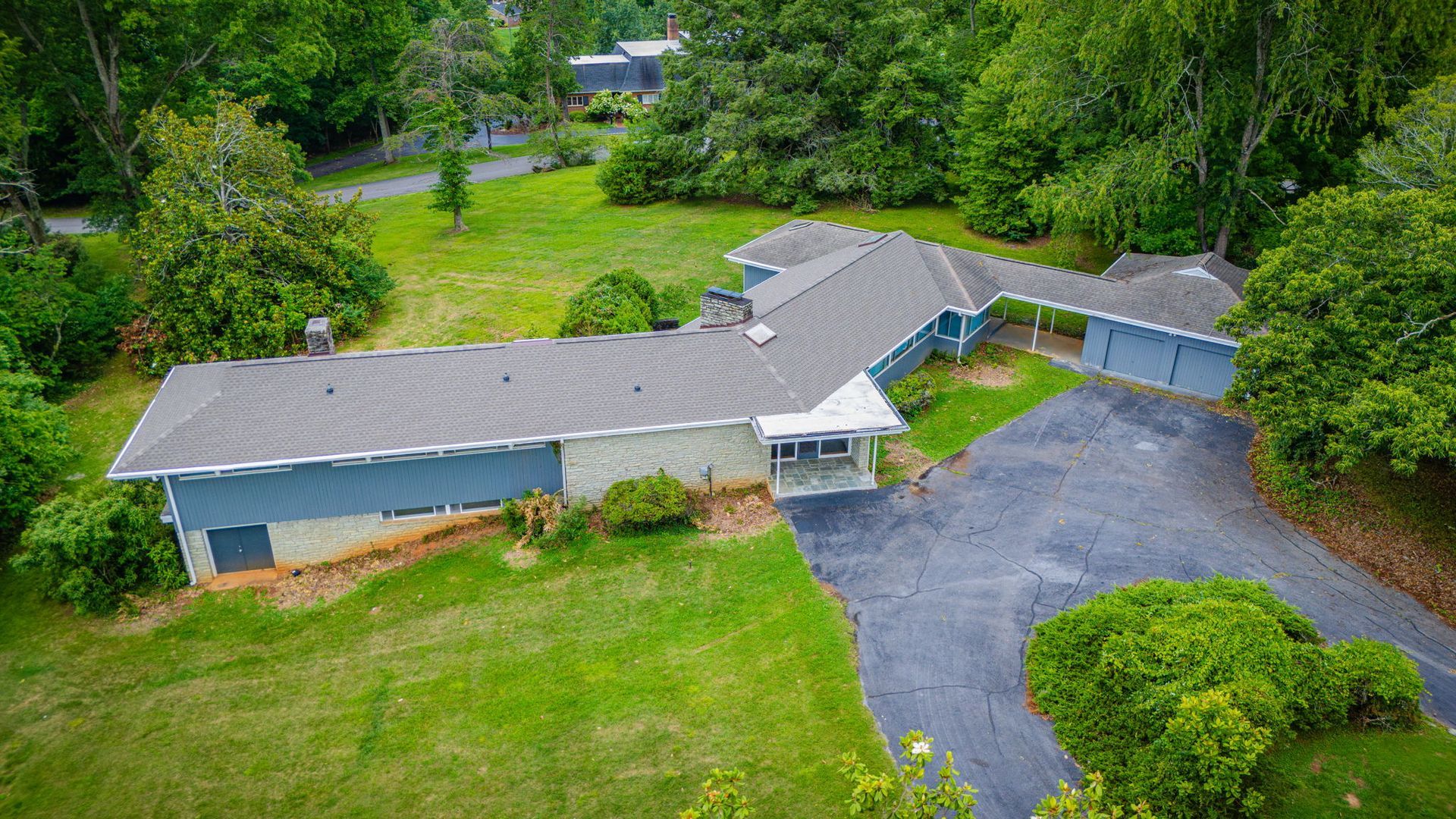 Aerial view of a long gray-roofed house with driveway, green lawn, and surrounding trees.