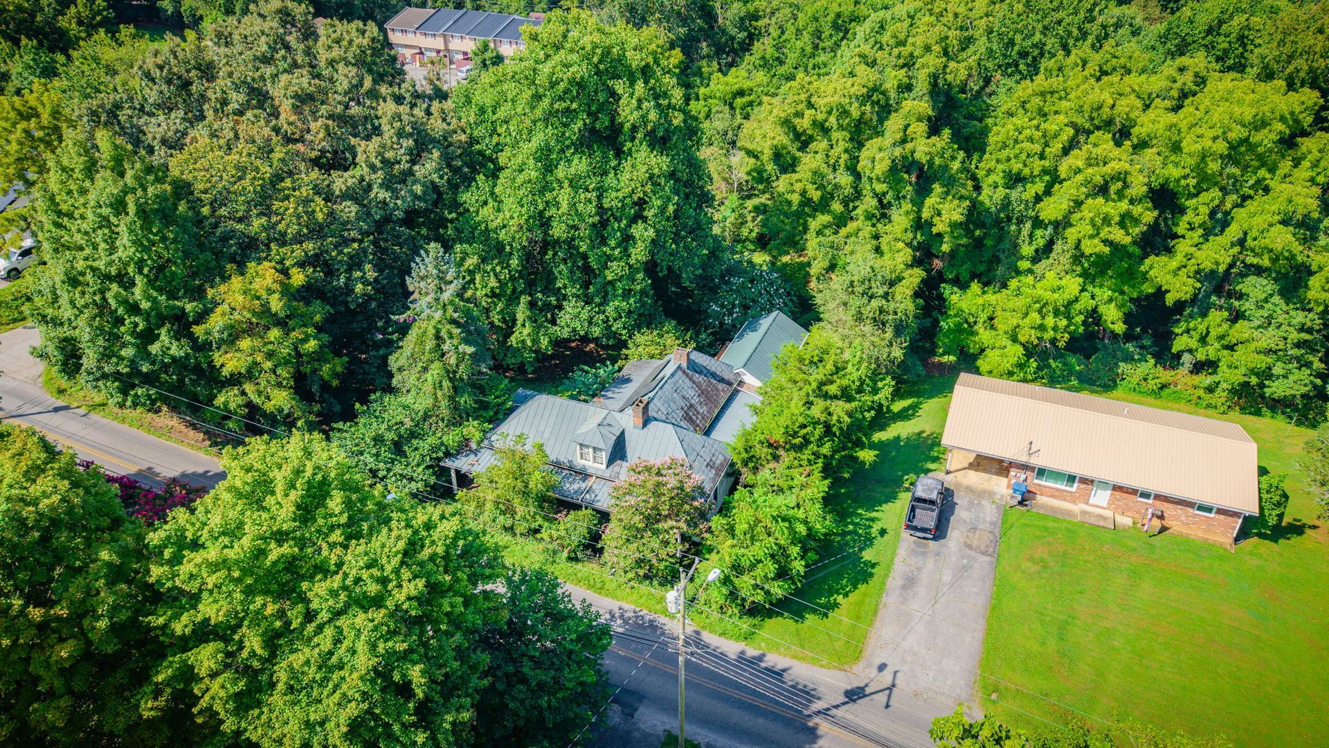 Aerial view of a house and outbuilding beside a road, surrounded by dense green trees and lawn.