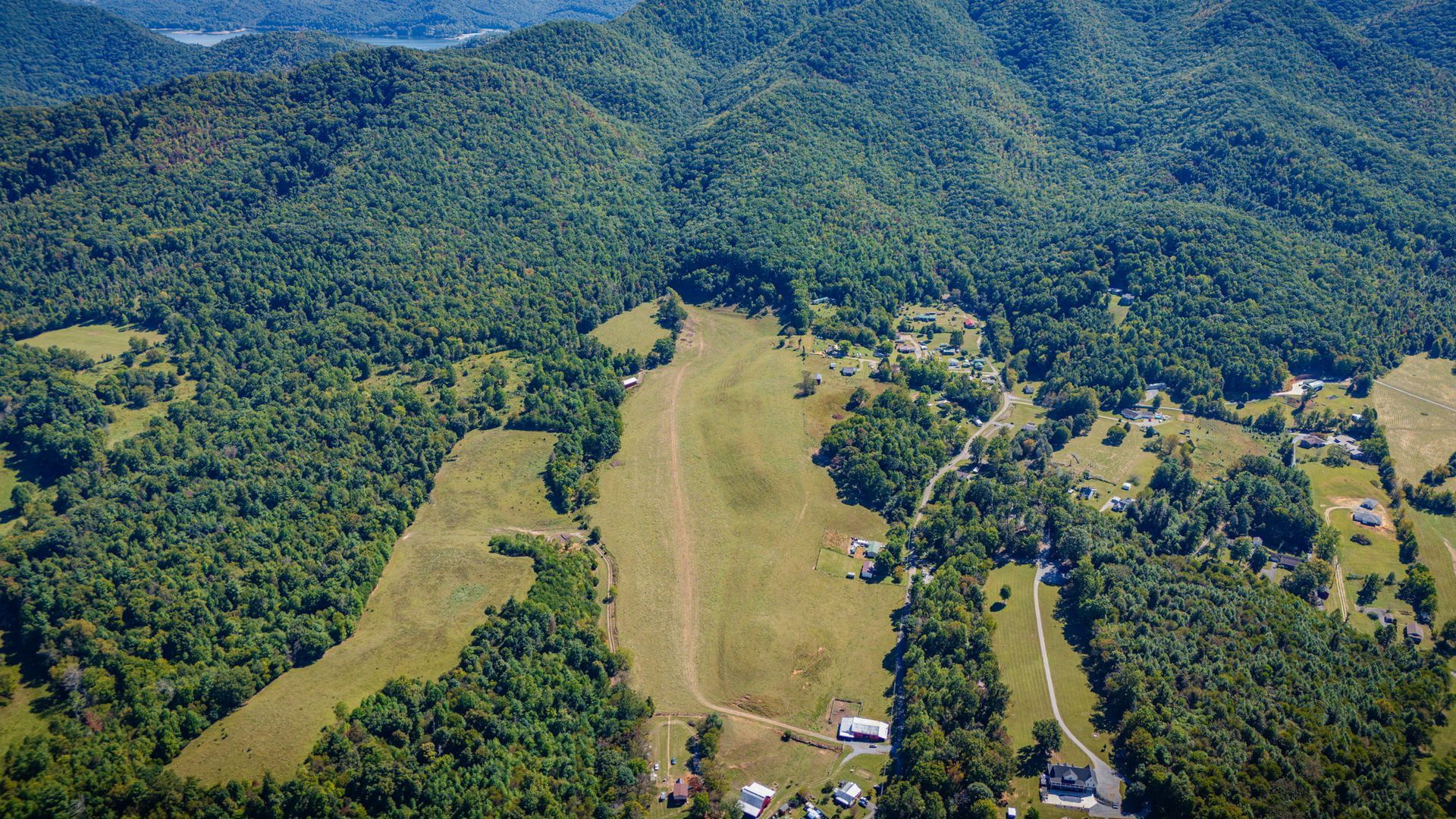 Aerial view of a grassy airstrip cutting through dense green forest in a mountainous valley.