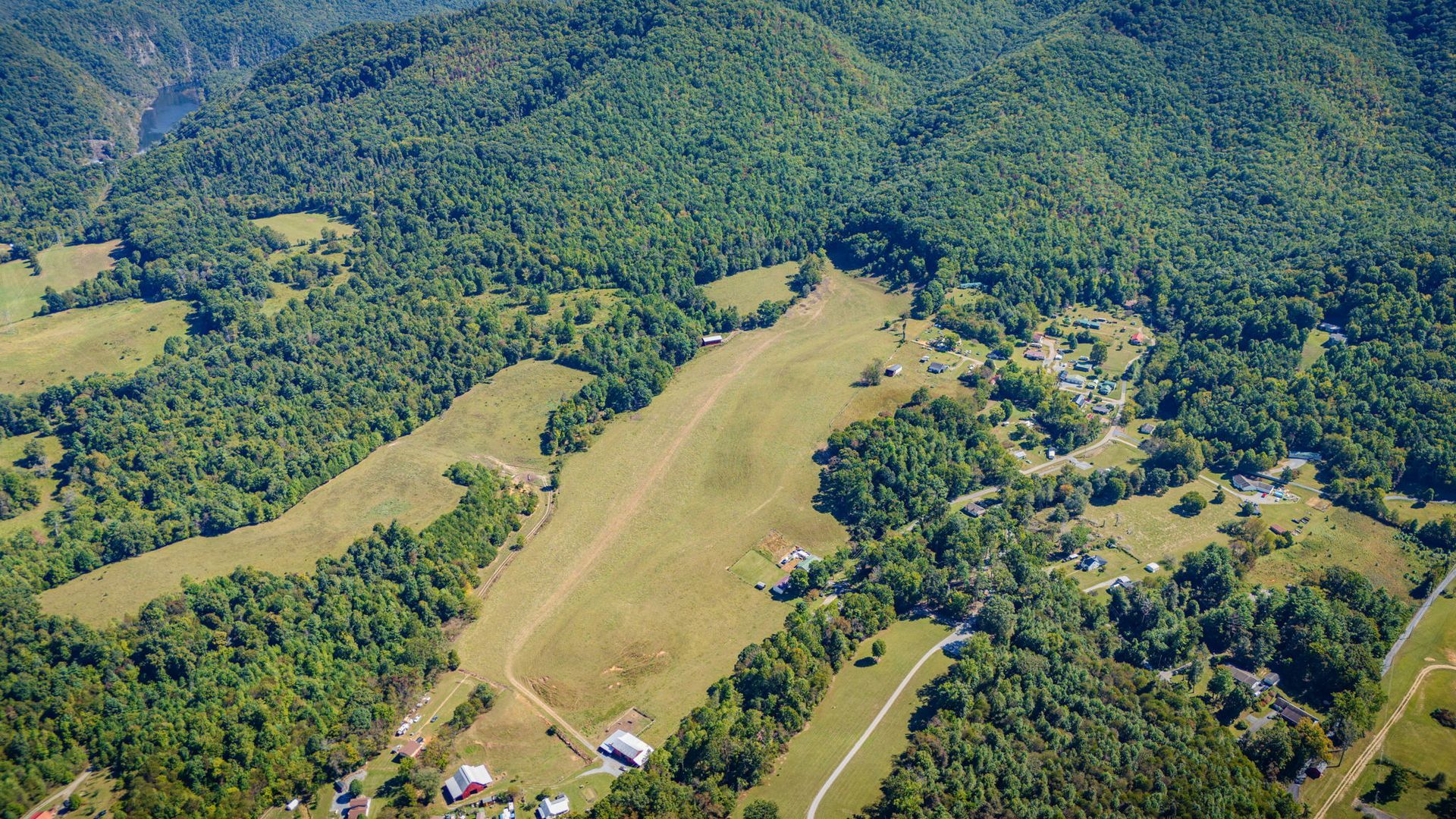 Aerial view of a grassy airstrip cut into dense green forest with a few small buildings nearby