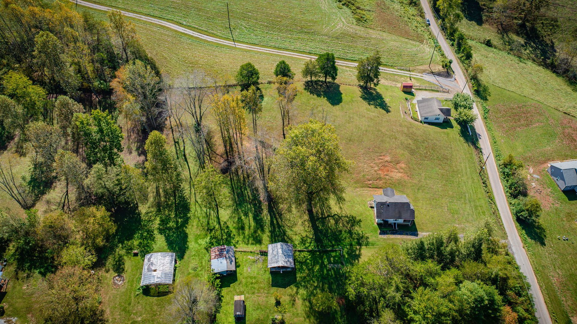 Aerial view of a rural green field with several small buildings, trees, and a dirt road along the edge