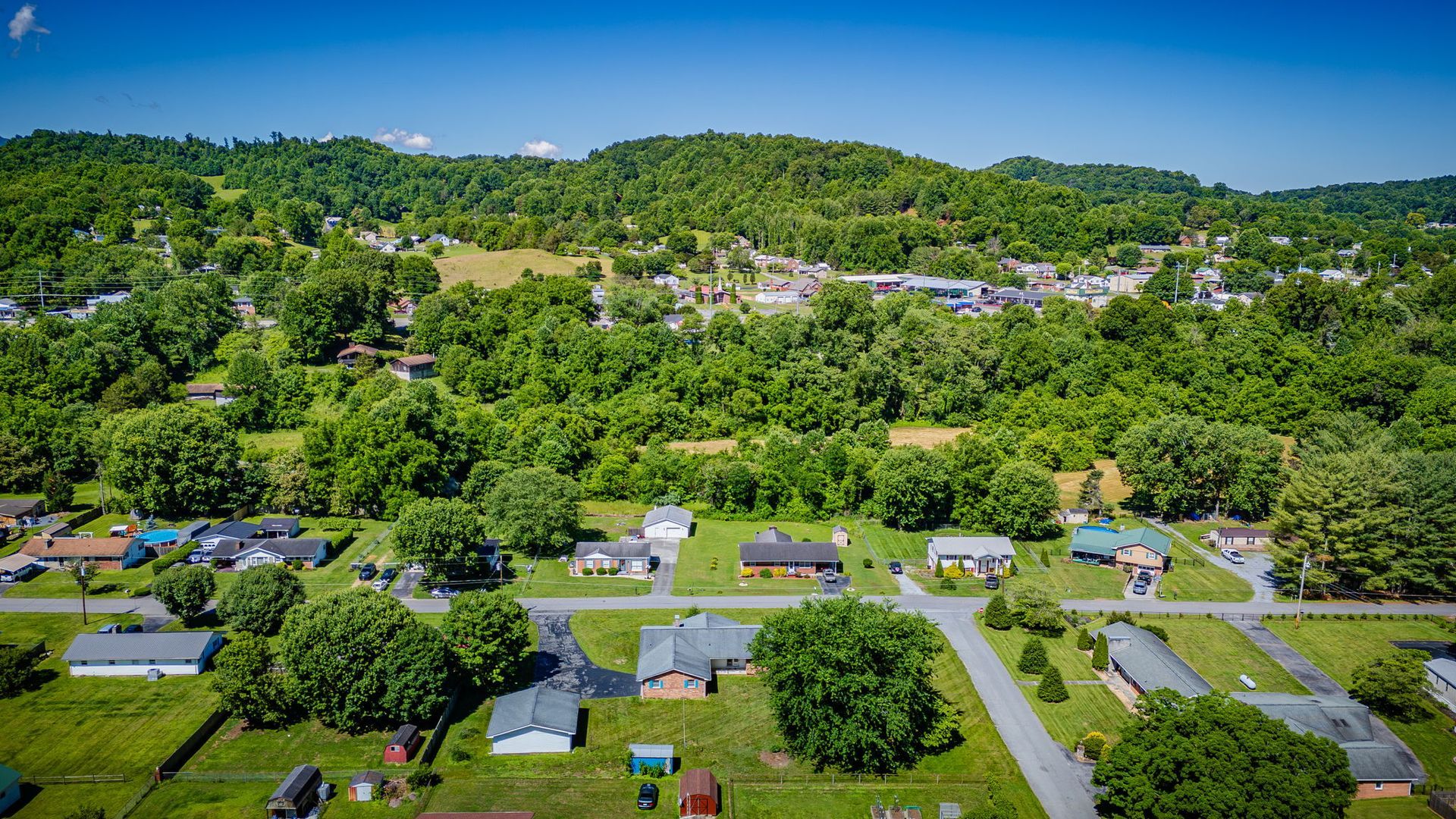 Aerial view of a green suburban neighborhood with houses, roads, and hills under a blue sky.