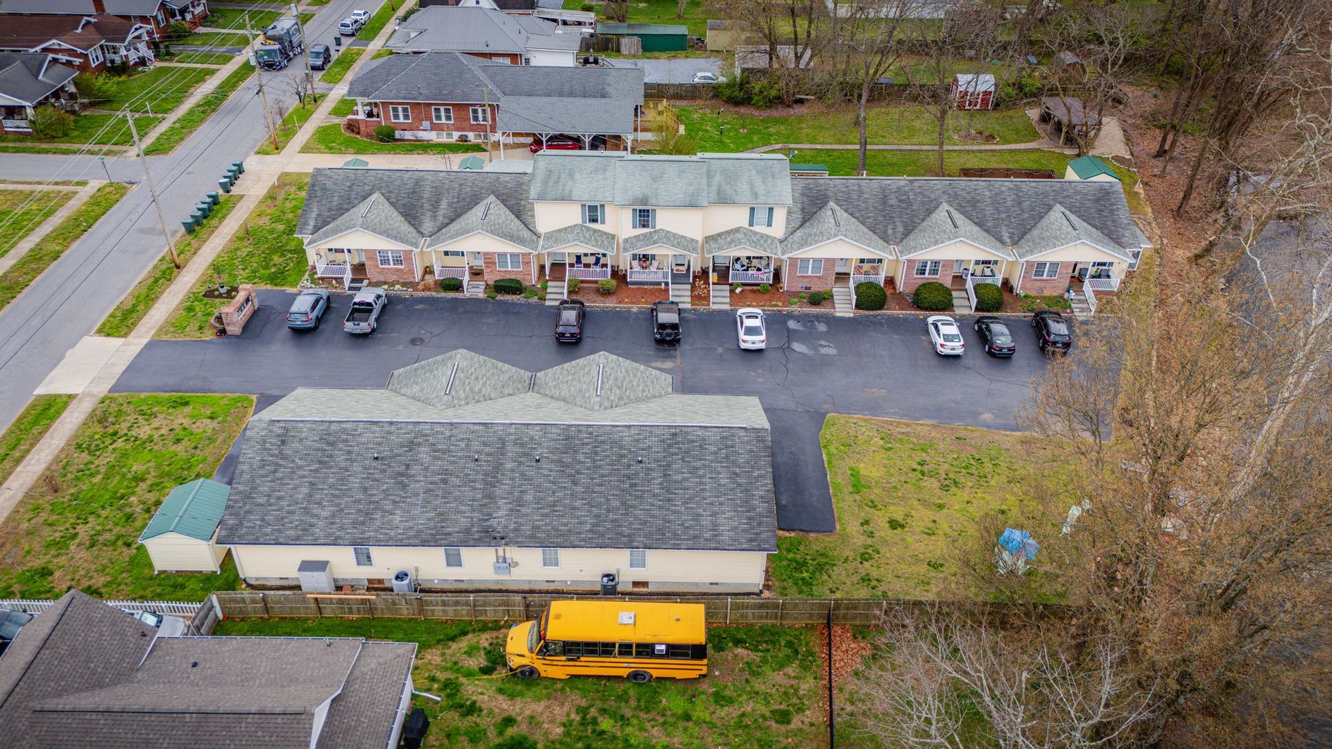 Aerial view of a long, single-story apartment complex with a paved parking lot and a yellow school bus parked nearby.