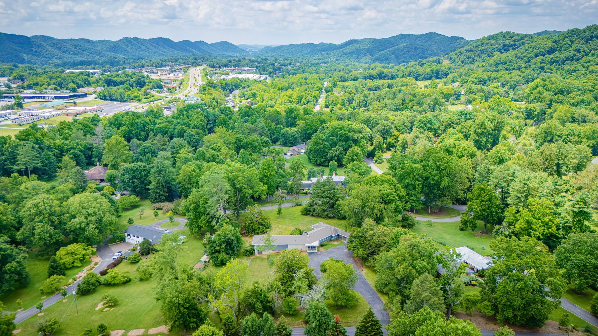 Aerial view of a lush green suburban neighborhood with scattered houses and wooded hills in the distance