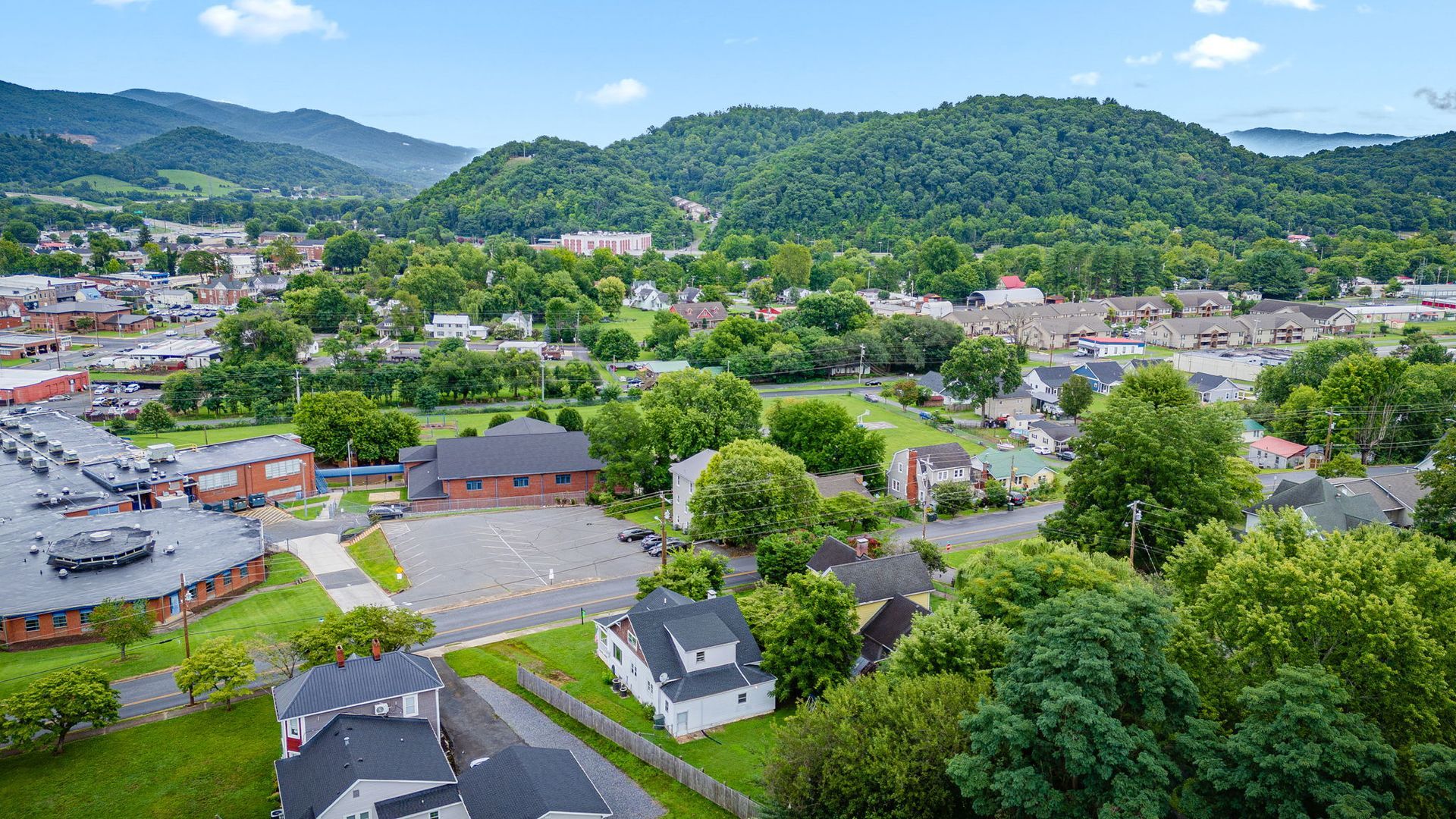 Aerial view of a small town with houses, roads, trees, and green hills under a blue sky.