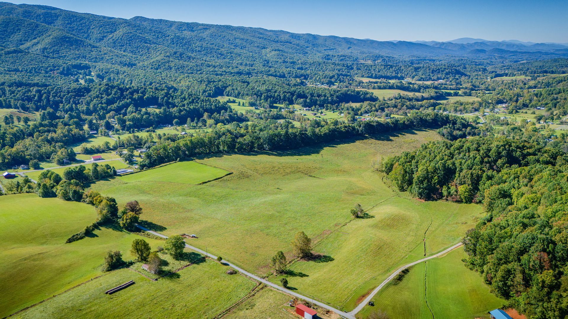 Aerial view of lush green fields, wooded hills, and distant mountains under a clear sky.