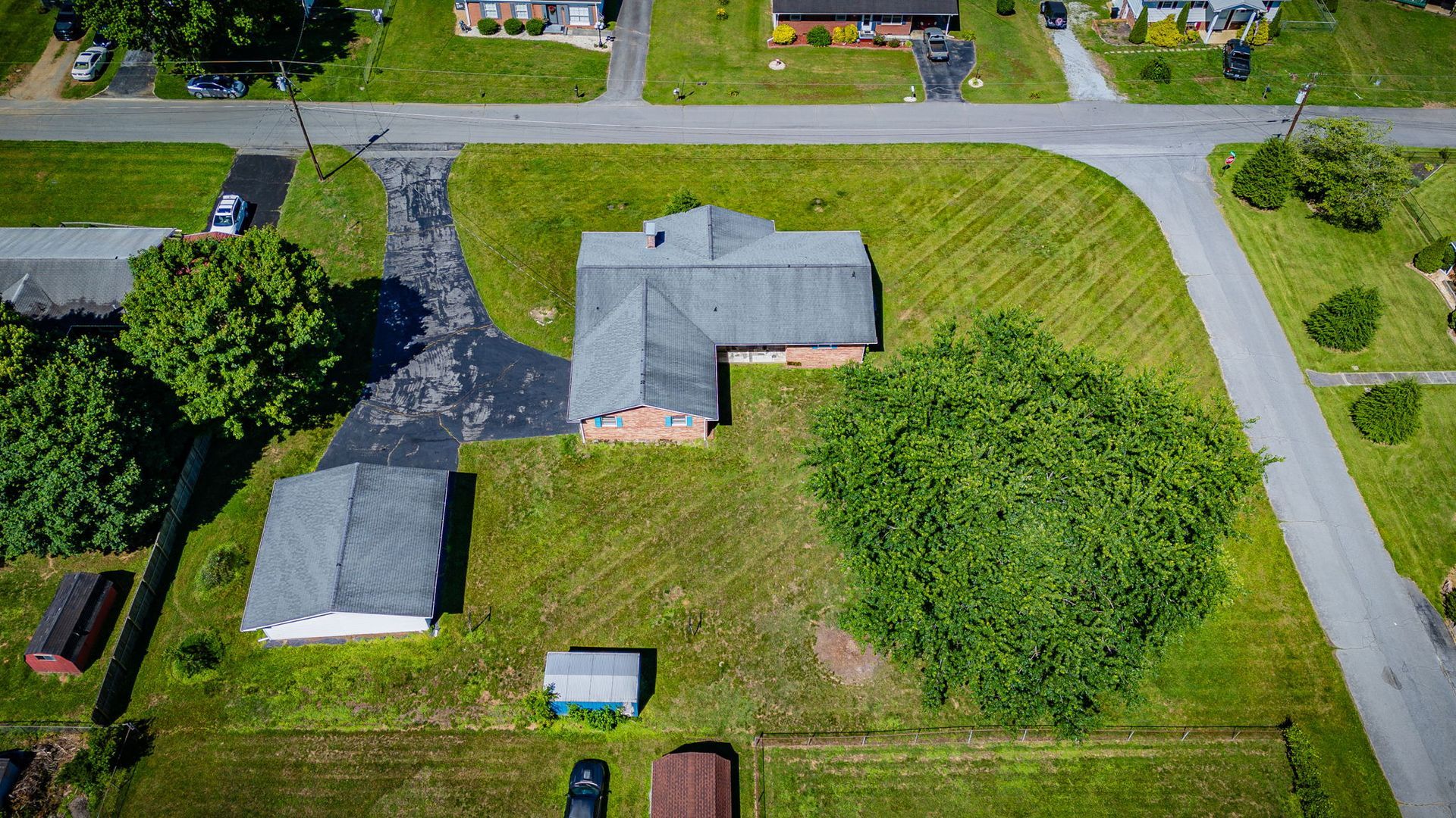 Aerial view of a suburban yard with a gray house, detached garage, trees, and surrounding roads