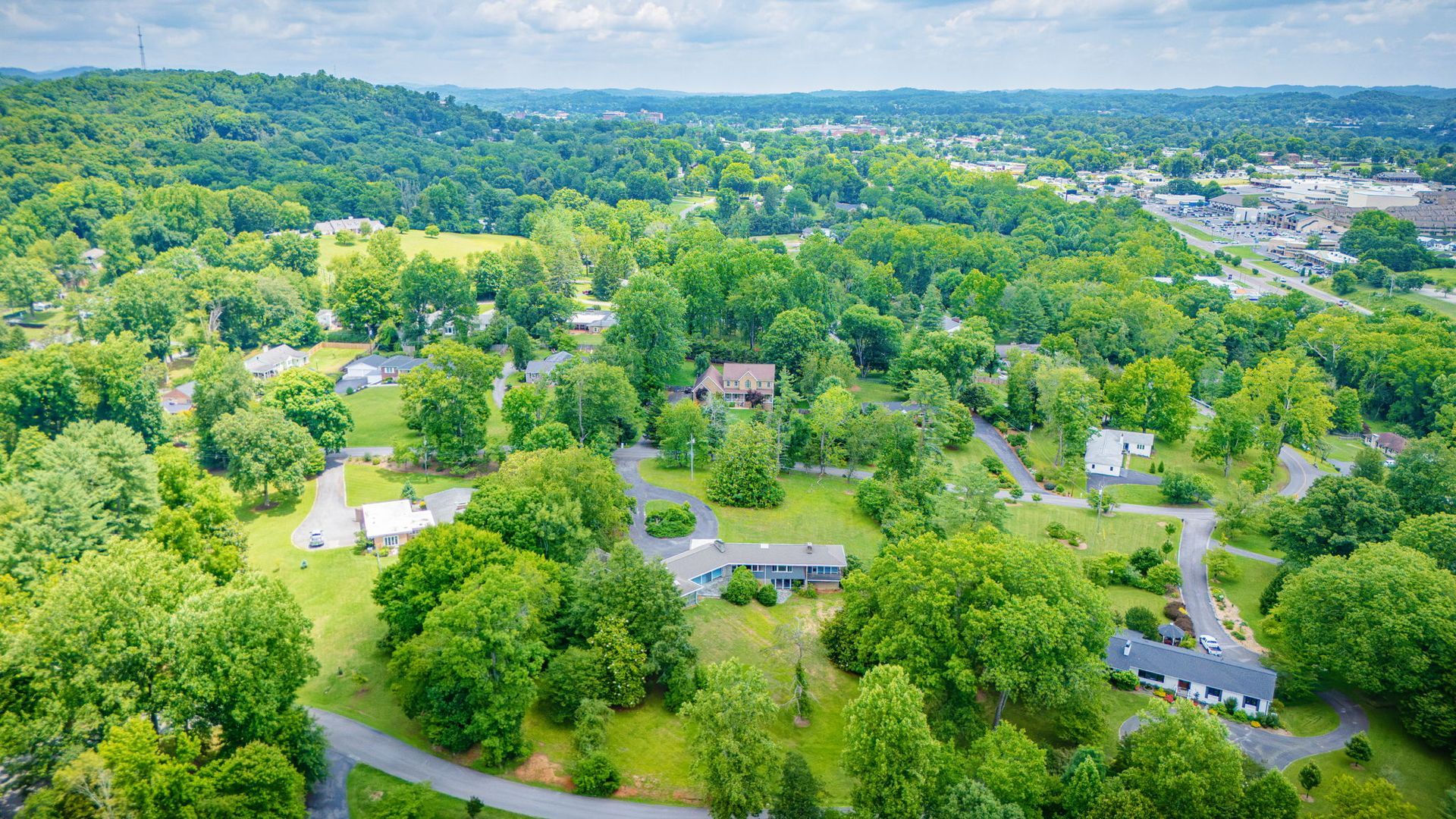 Aerial view of a leafy suburban neighborhood with winding roads, houses, and rolling green hills.