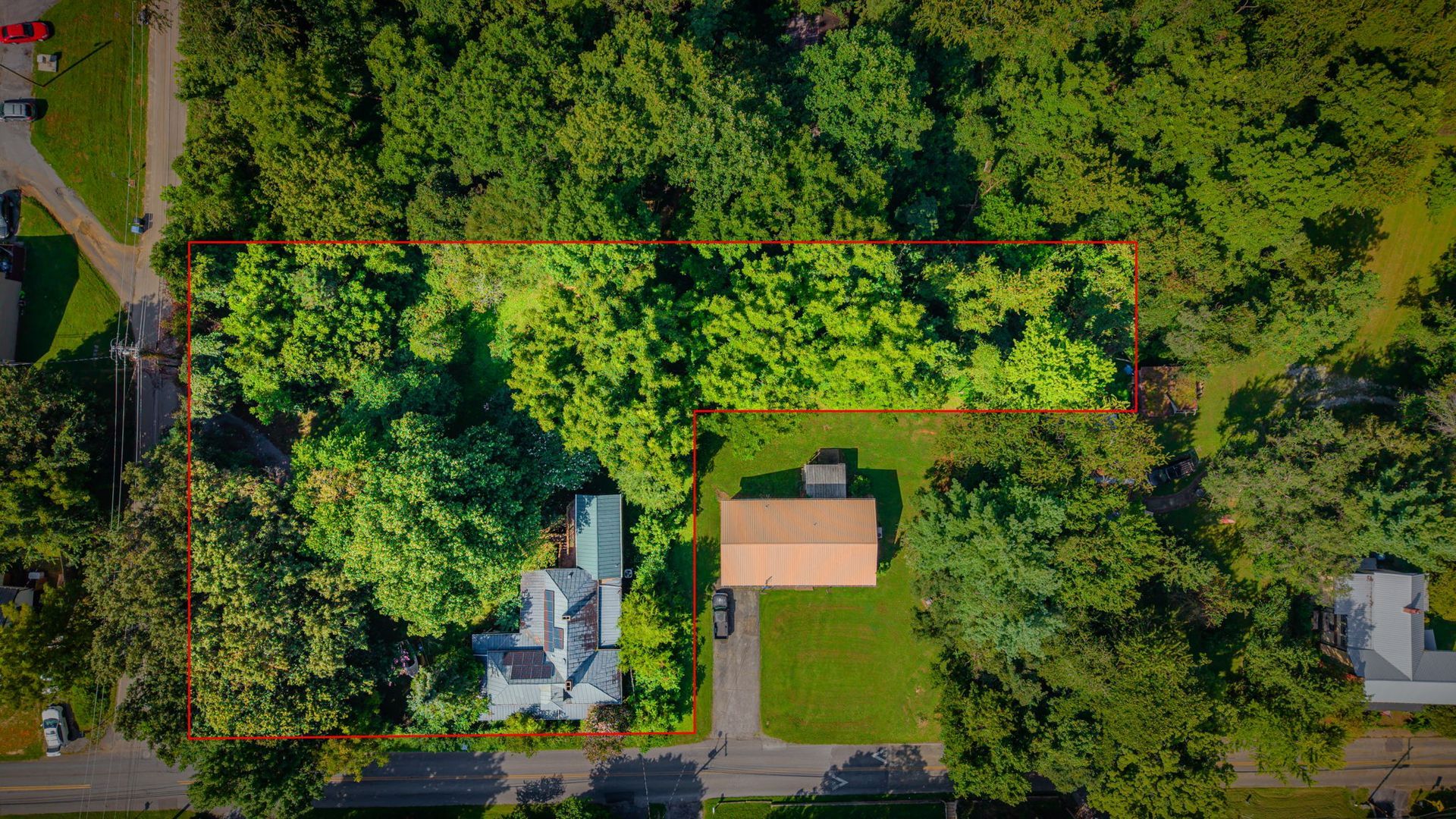 Aerial view of a wooded residential street with houses and a parked car, crossed by a red dashed boundary line.