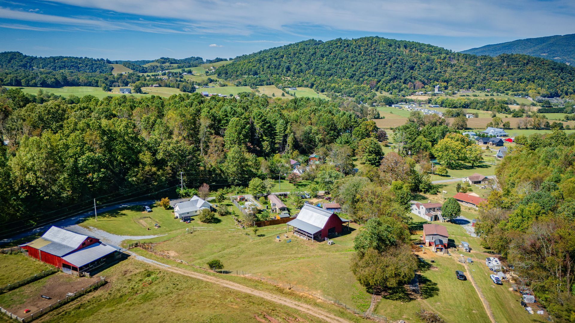 Aerial view of a rural farm with red-roofed buildings, green fields, and forested hills under a clear sky