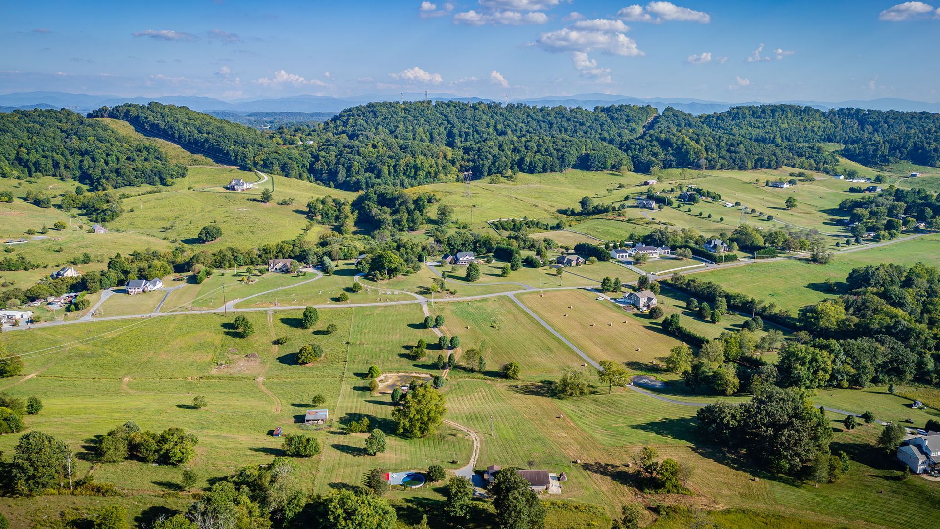 Wide green countryside with rolling hills, scattered houses, and a forested ridge under a blue sky
