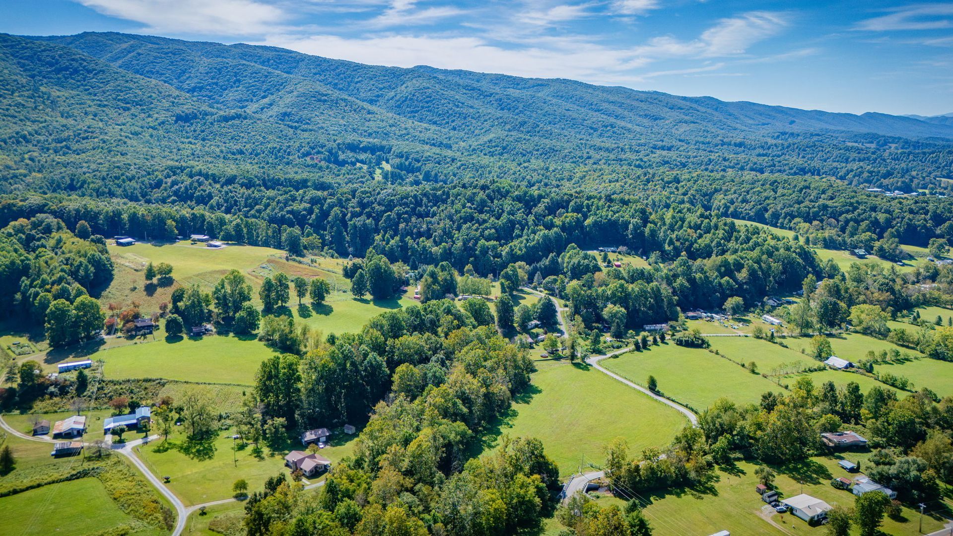Sunny rural valley with green fields, scattered houses, and forested mountains in the distance
