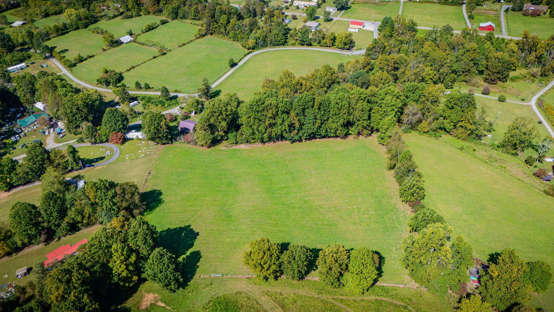 Aerial view of a green rural field bordered by trees, roads, and scattered houses.