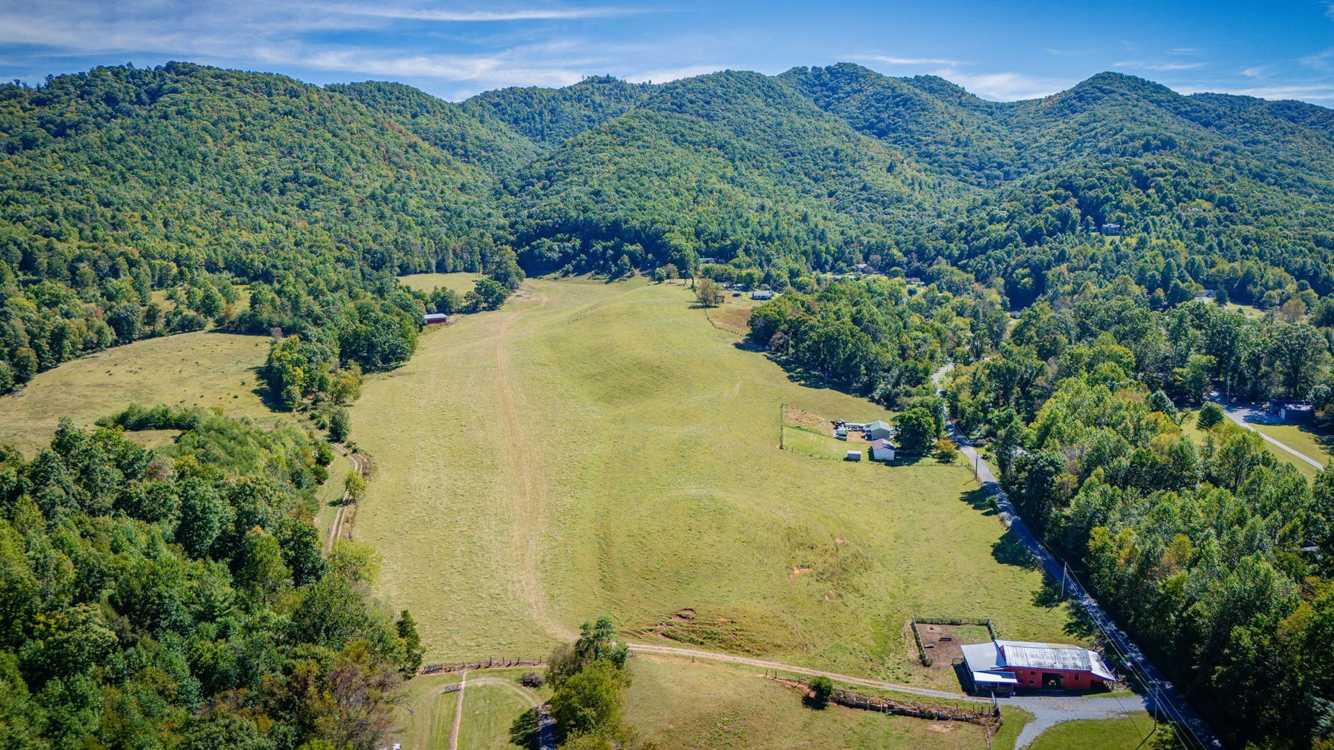 Aerial view of a green valley meadow surrounded by forested hills and mountains, with a few small buildings