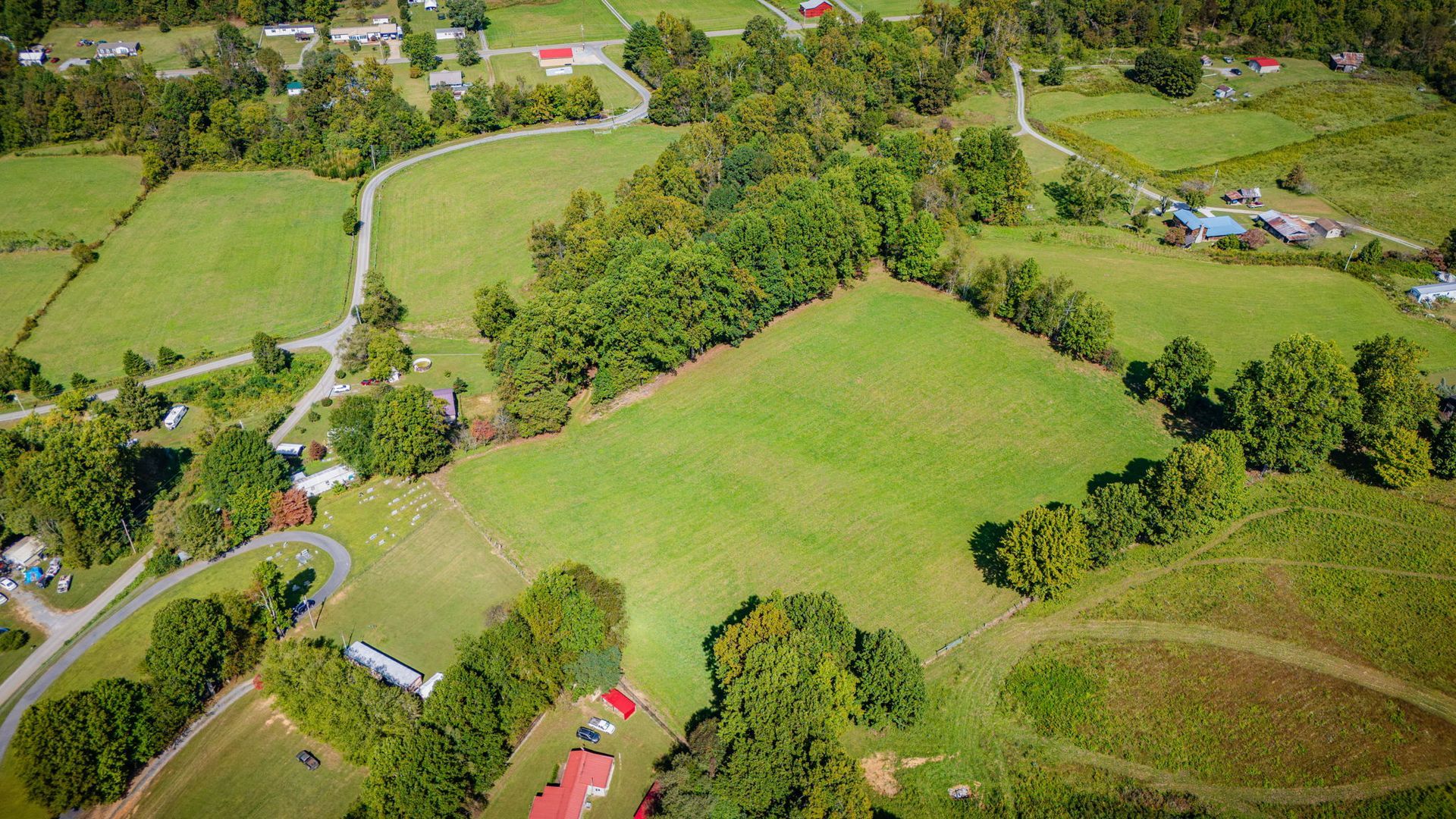 Aerial view of green fields, winding roads, and scattered trees with a small red-roofed house.