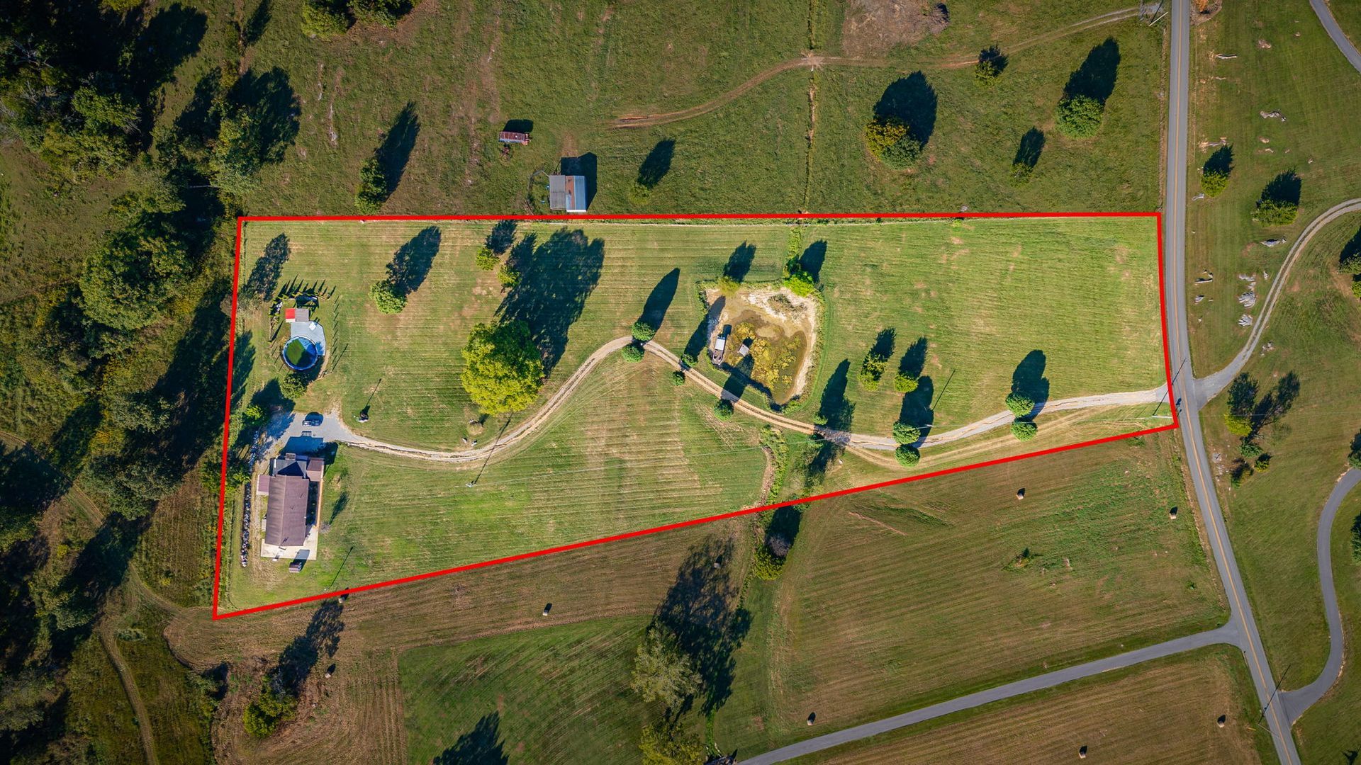 Aerial view of a fenced rural lot with trees, a small building, and a winding dirt path beside a road.