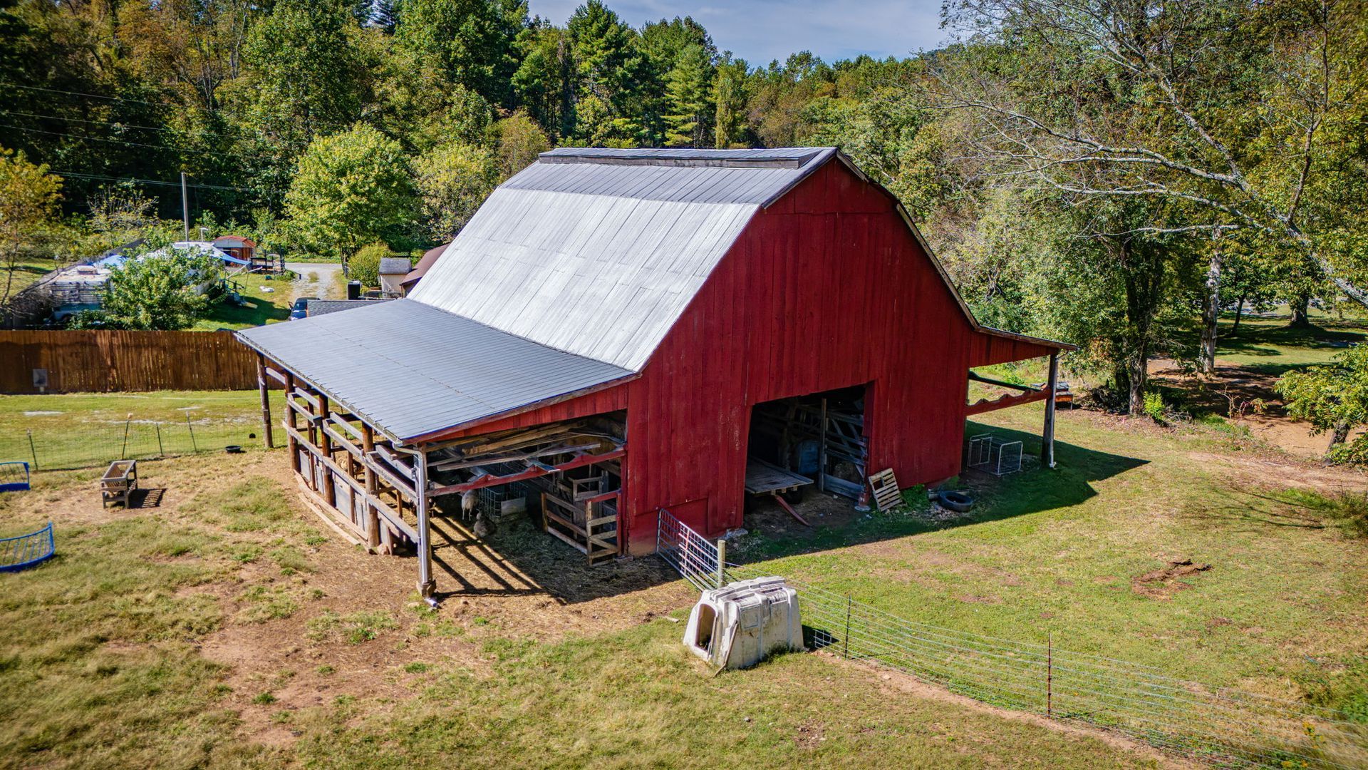 Red barn with silver roof in a grassy field, surrounded by trees and a wooden fence