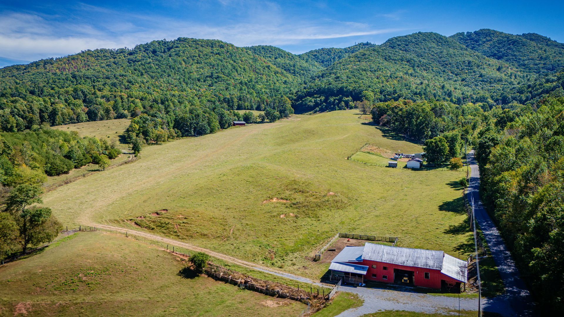 Aerial view of a green pasture and red barn beside a road, with forested hills in the background