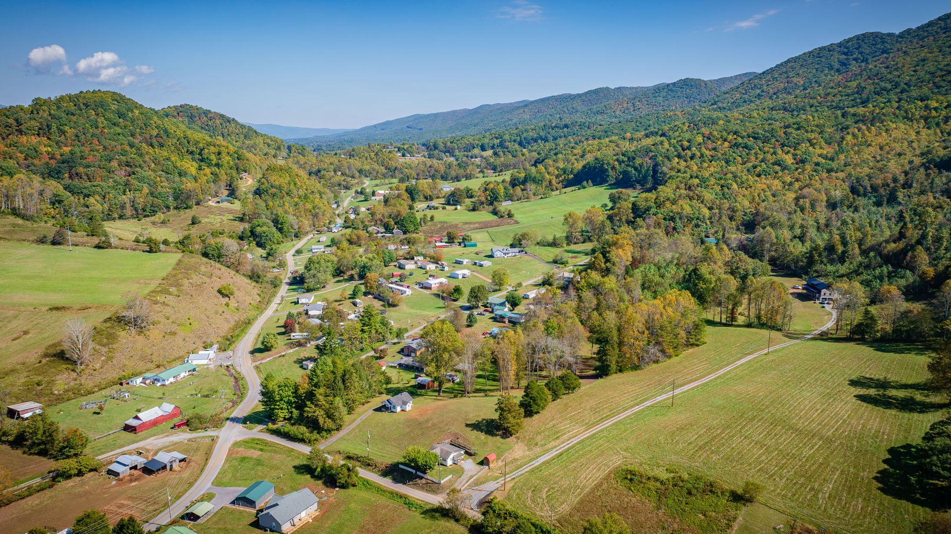 Aerial view of a rural valley with scattered houses, winding road, green fields, and forested hills under blue sky