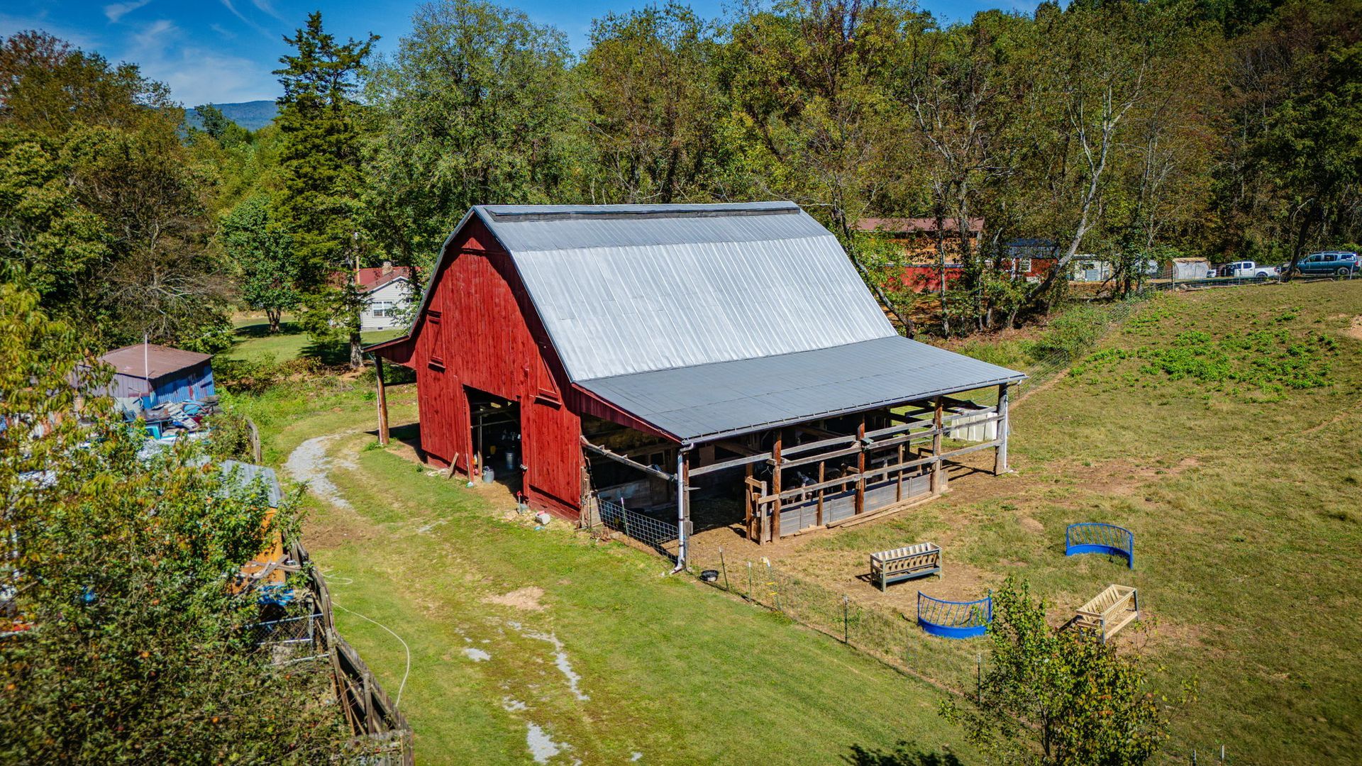 Red barn with gray roof in a grassy rural yard, surrounded by trees and outbuildings.