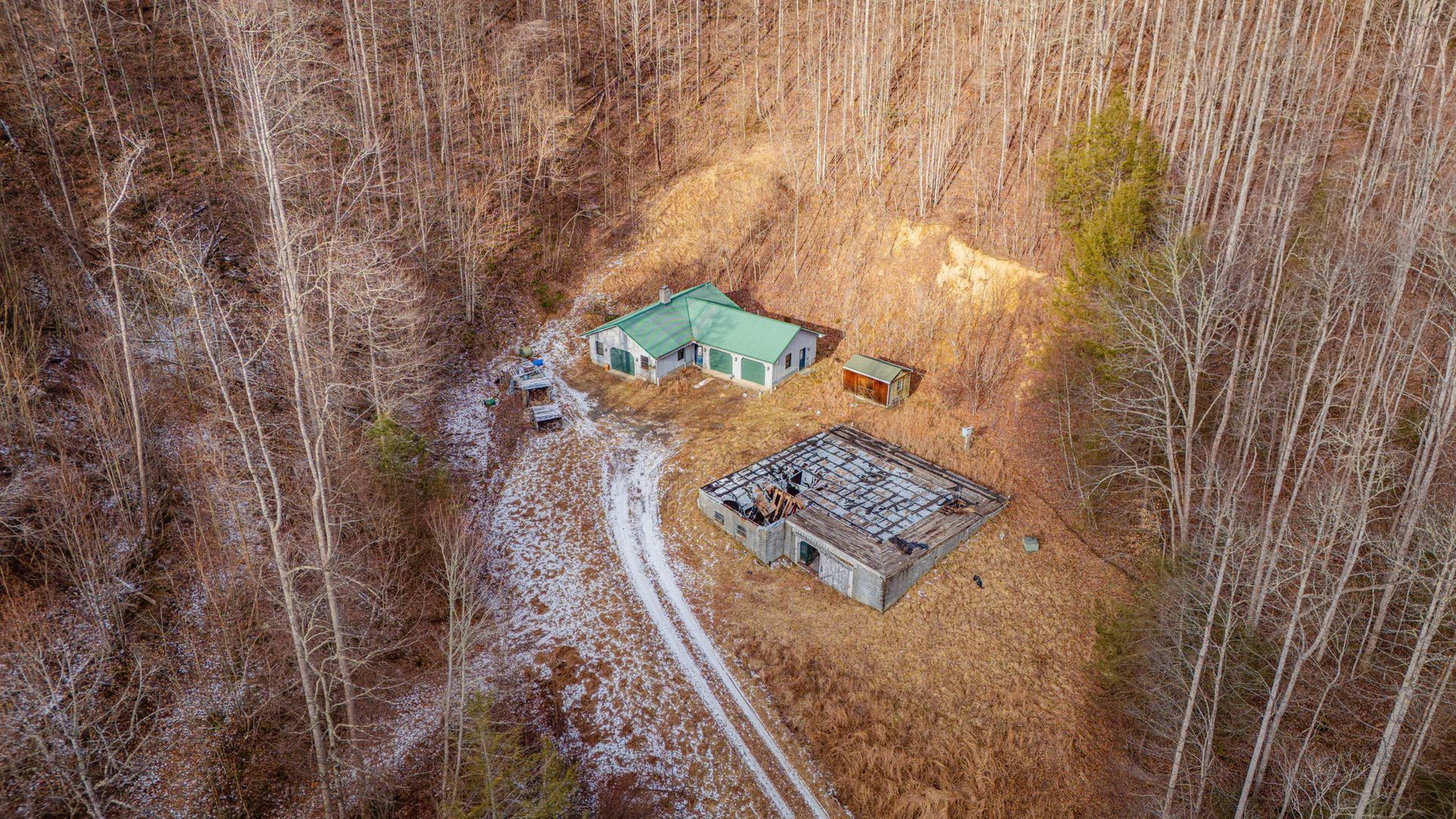 Aerial view of a home and outbuilding in a wooded, brown landscape with a snow-covered dirt road.