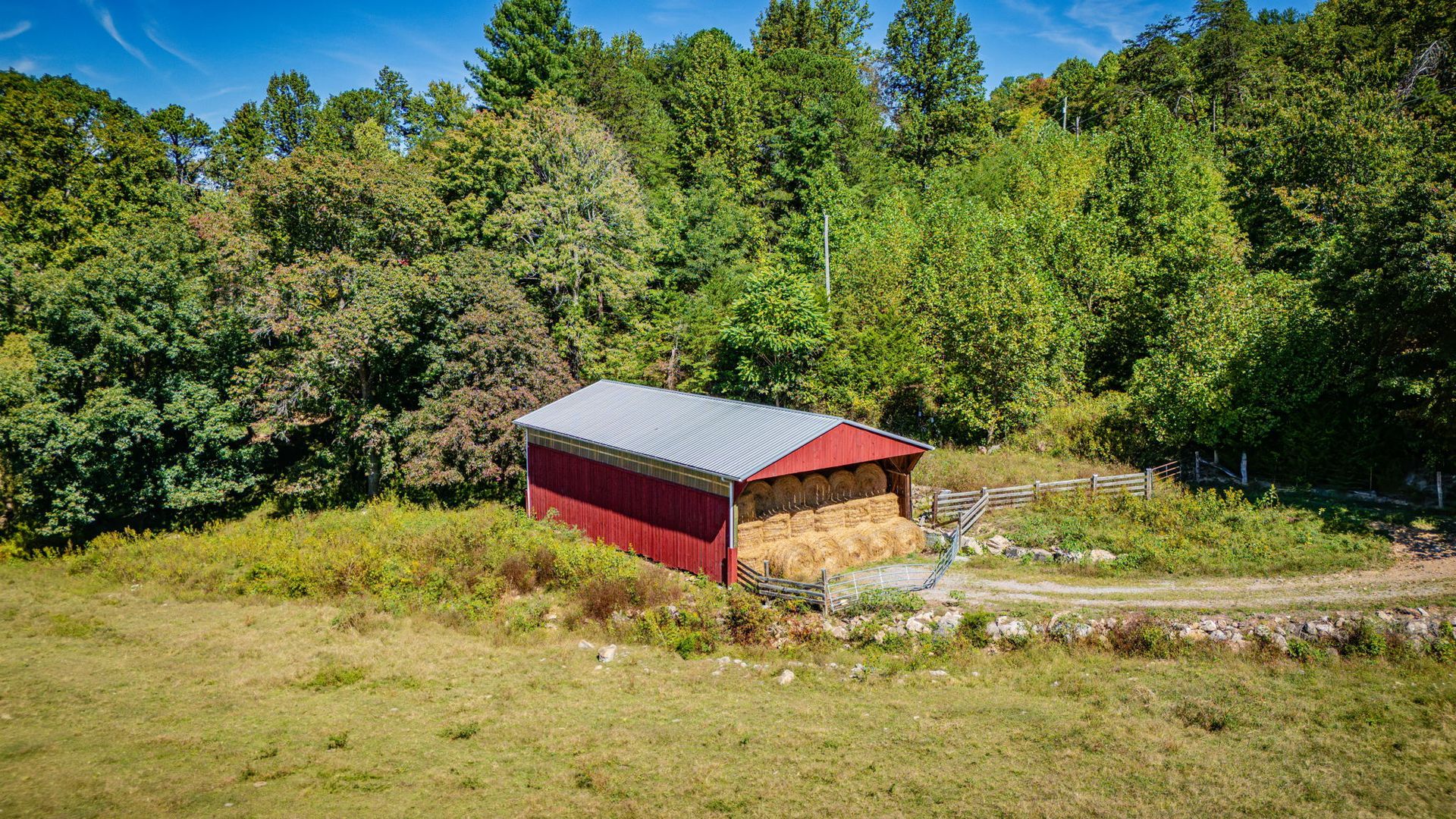Red barn beside a stone wall in a grassy field, surrounded by dense green trees