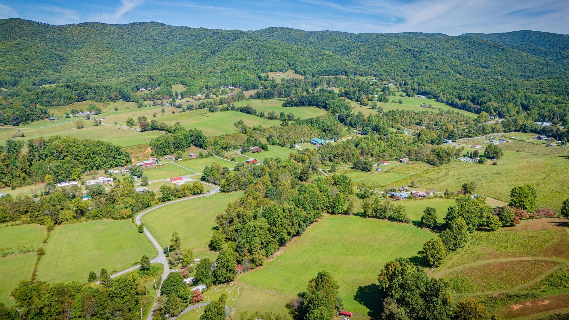 Aerial view of green farmland and scattered houses beside forested hills under a clear sky